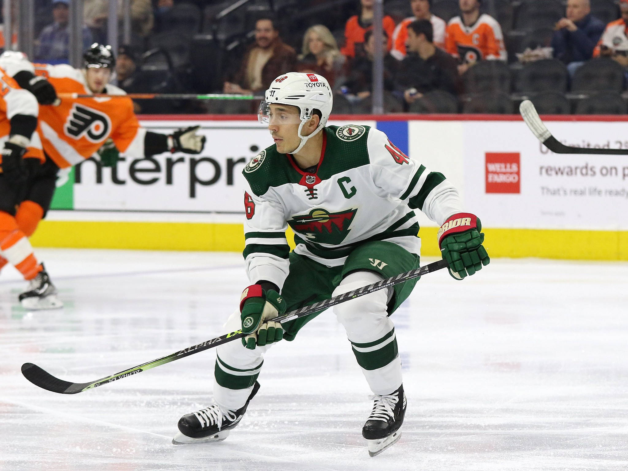 hockey 桌布 壁紙 — 一名專注的曲棍球運動員穿著綠色和白色的裝備，在明亮的冰場上帶著球桿滑行比賽中。