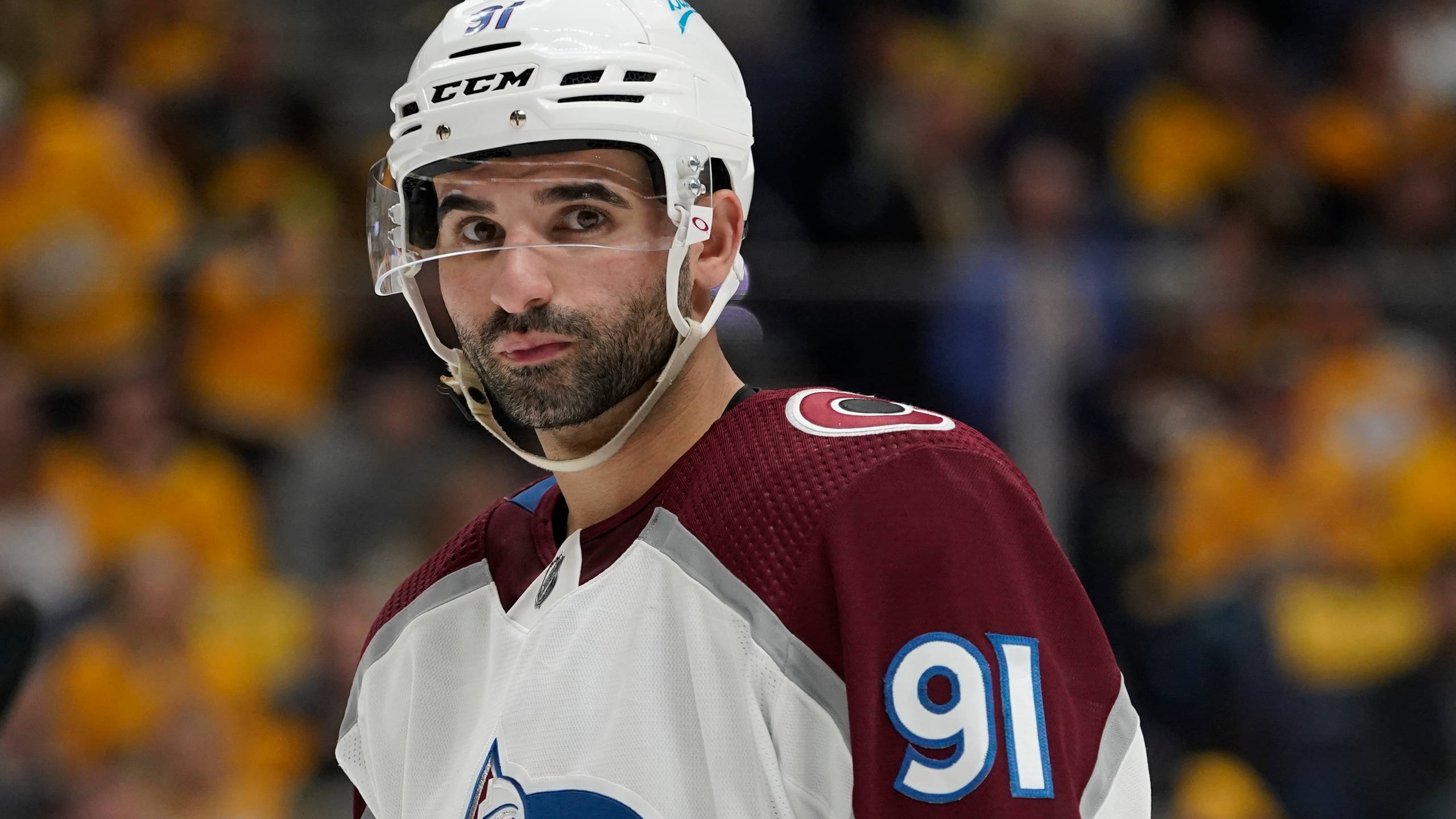 nazem kadri 桌布 壁紙 — 一名專注的曲棍球運動員身穿棗紅色和白色球衣，號碼91，在比賽中凝視著，背景是模糊的黃色人群。