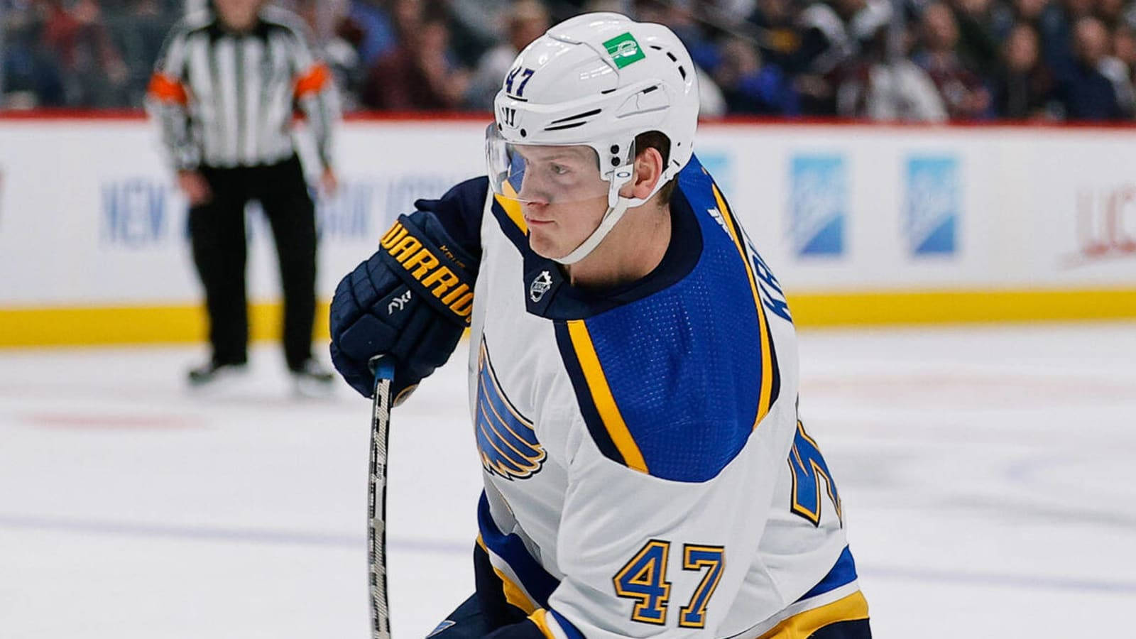 St Louis Blues 桌布 壁紙 — 一名專注的曲棍球運動員身穿白色、藍色和金色球衣，背號47，在冰上滑行，背景有裁判。