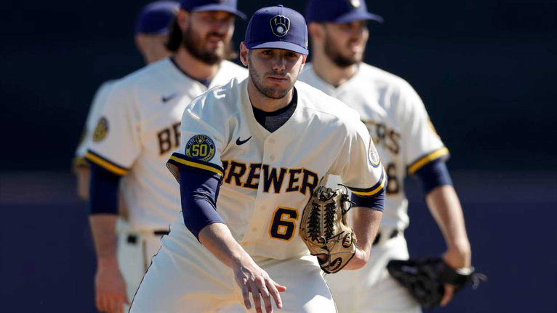 Brewers 桌布 壁紙 — 一名專注的棒球運動員身穿釀酒人隊制服蹲下準備防守，在陽光明媚的日間比賽中，背景的隊友模糊不清。