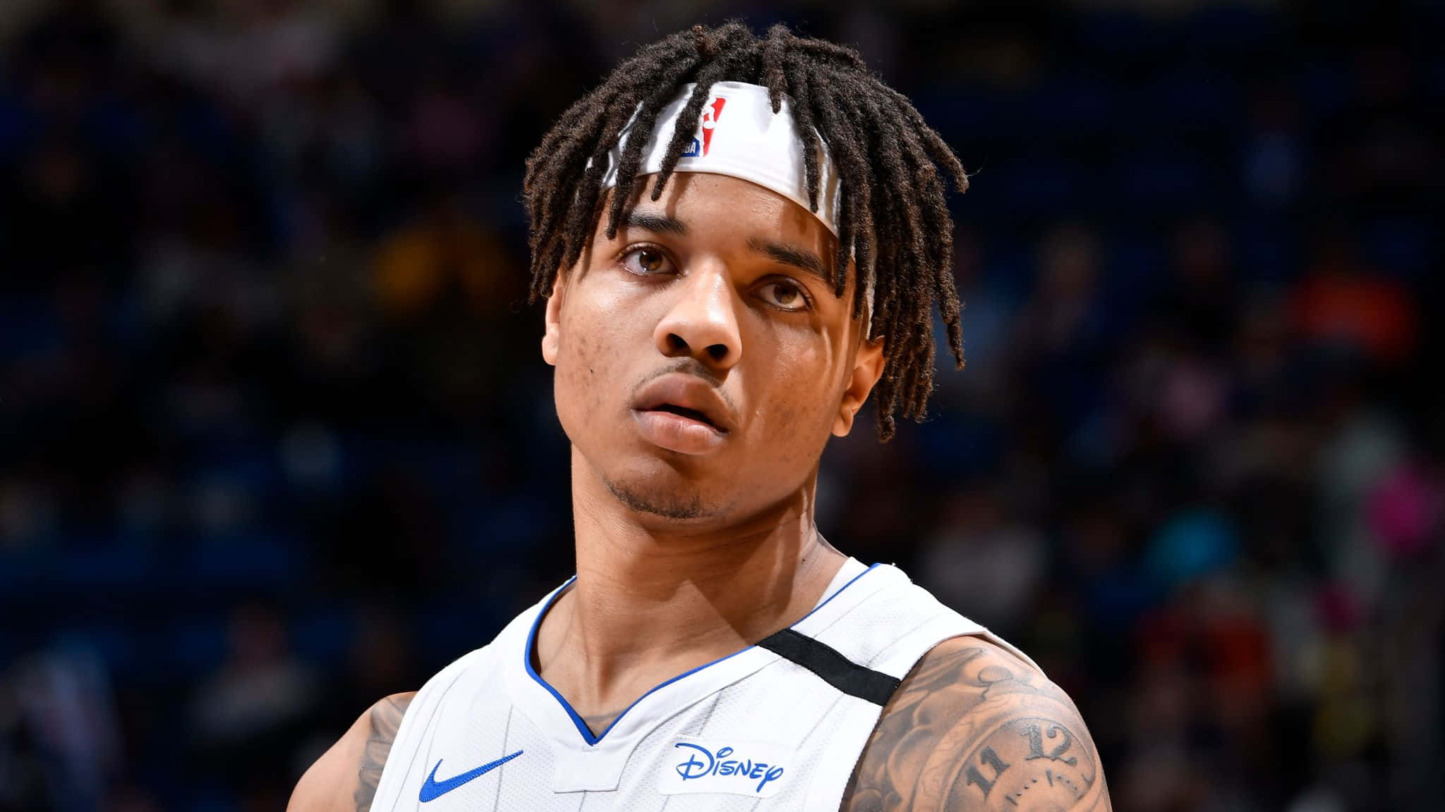 NBA 桌布 壁紙 — 一名專注的籃球運動員，留著辮子頭，戴著白色頭帶，在場館燈光下比賽時凝視著前方。
