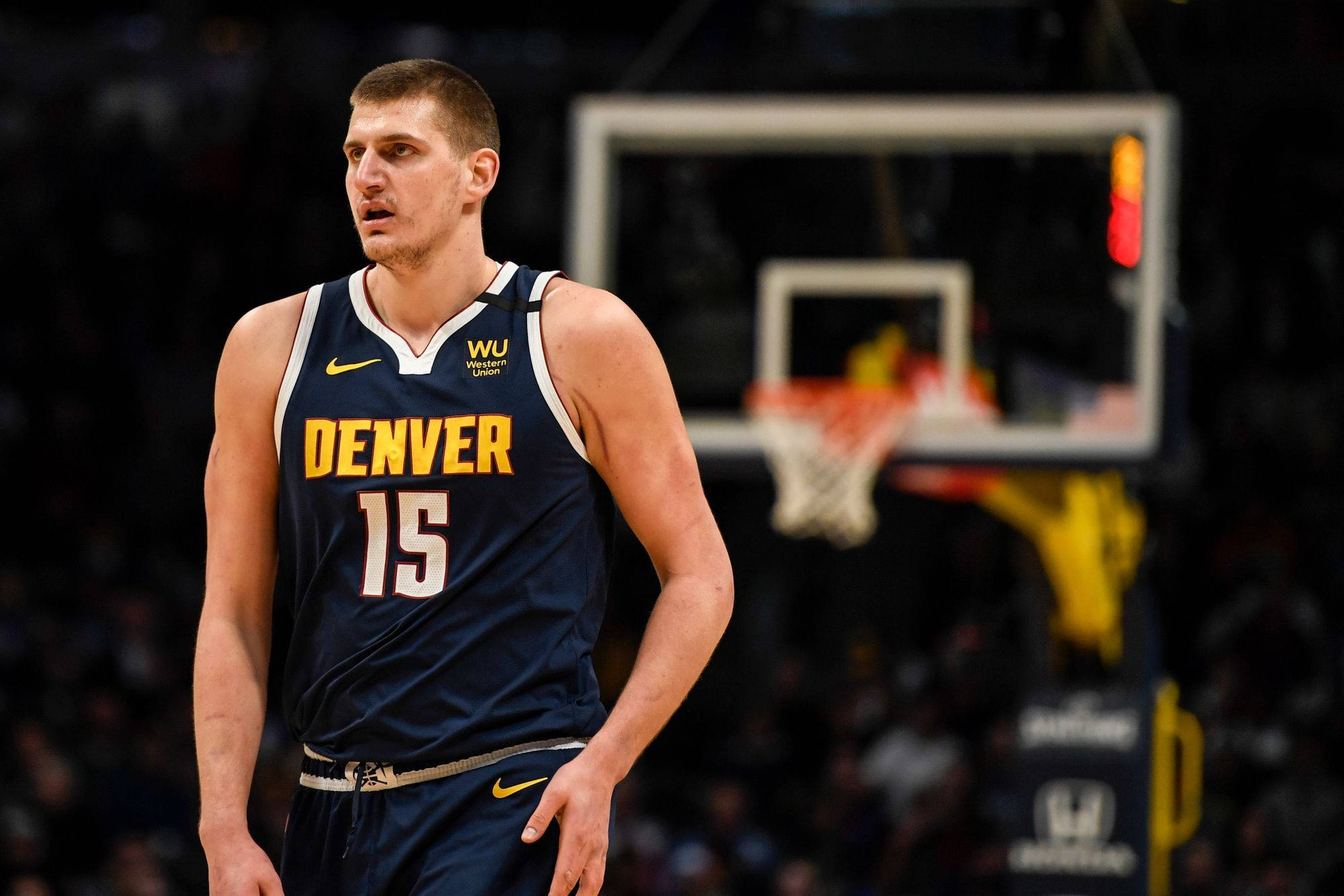 Jokic 桌布 壁紙 — 一名專注的籃球運動員身穿丹佛球衣，在場館燈光下站在球場上，身後籃框模糊。