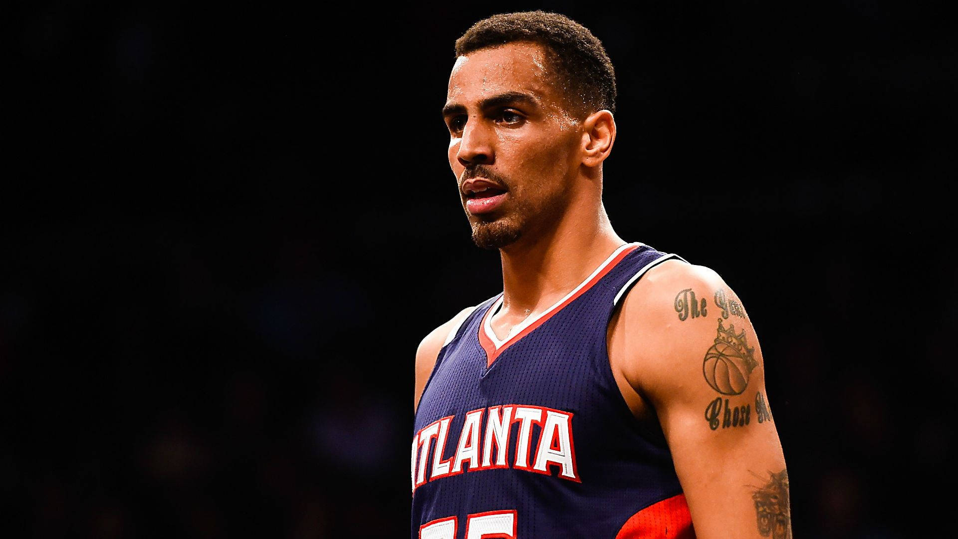 Atlanta Hawks 桌布 壁紙 — 一名專注的籃球運動員身穿亞特蘭大球衣，在黑暗的競技場背景下凝視著。