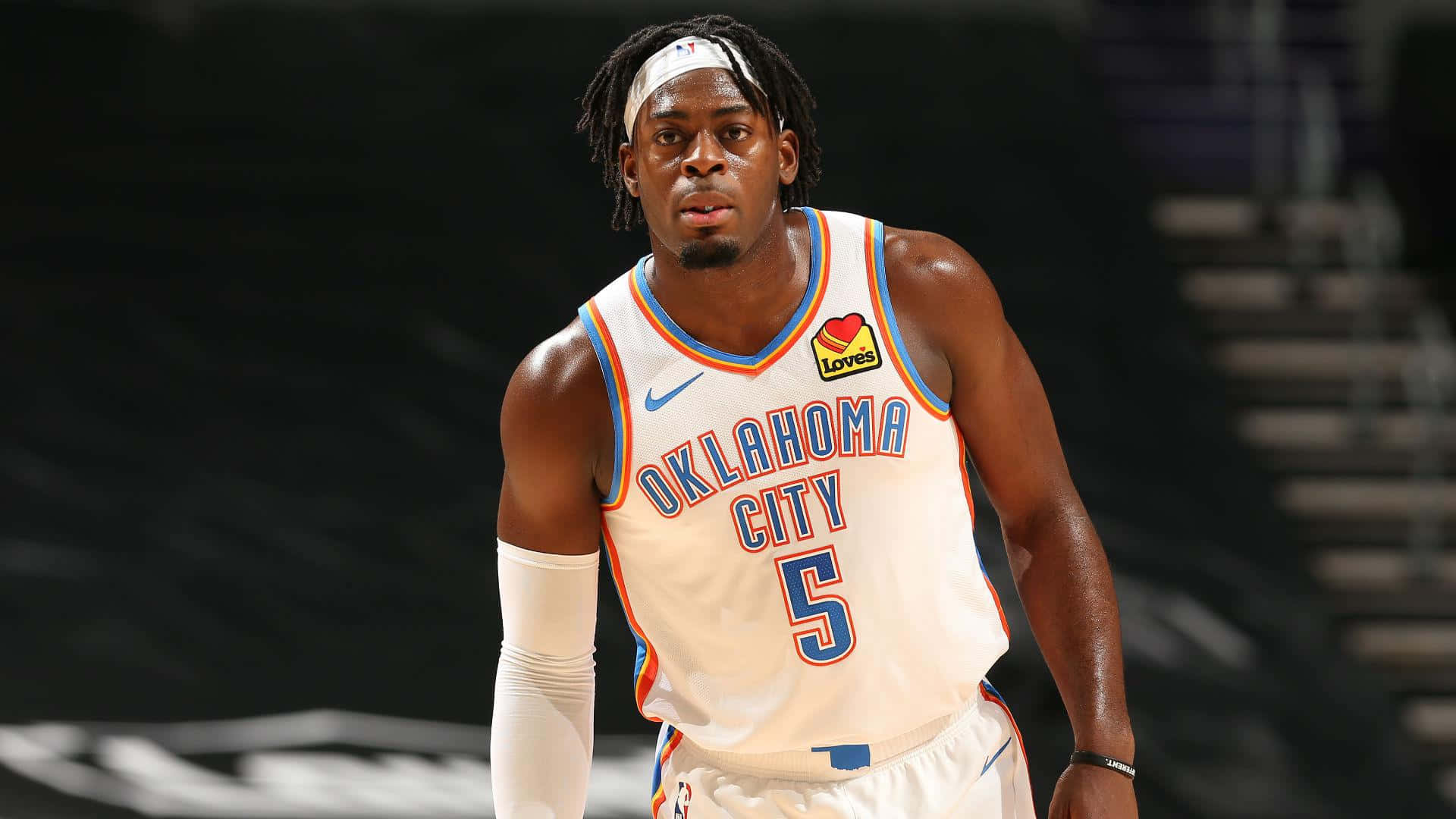 NBA 桌布 壁紙 — 一名專注的籃球運動員身穿奧克拉荷馬城球衣，在燈光昏暗的球場上準備就緒，身後是模糊的體育館座位。