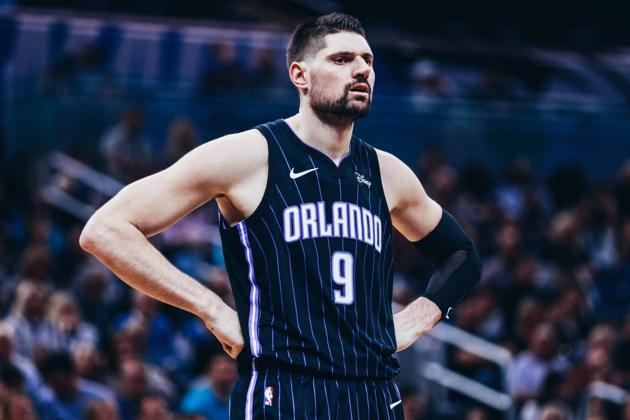 NBA 桌布 壁紙 — 一名專注的籃球運動員身穿奧蘭多球衣，在明亮的場館燈光下比賽期間雙手叉腰站立。