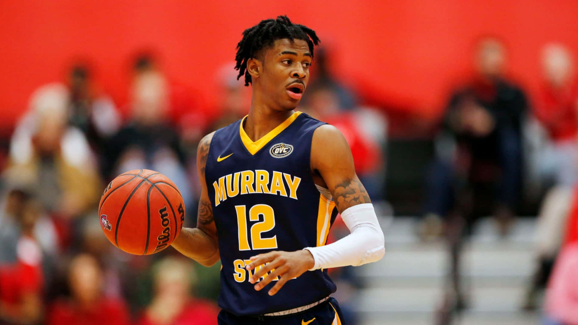 Ja Morant 動圖 桌布 壁紙 — 一名專注的籃球運動員身穿海軍藍 Murray State 球衣，在模糊的紅色人群背景下運著橙色籃球。