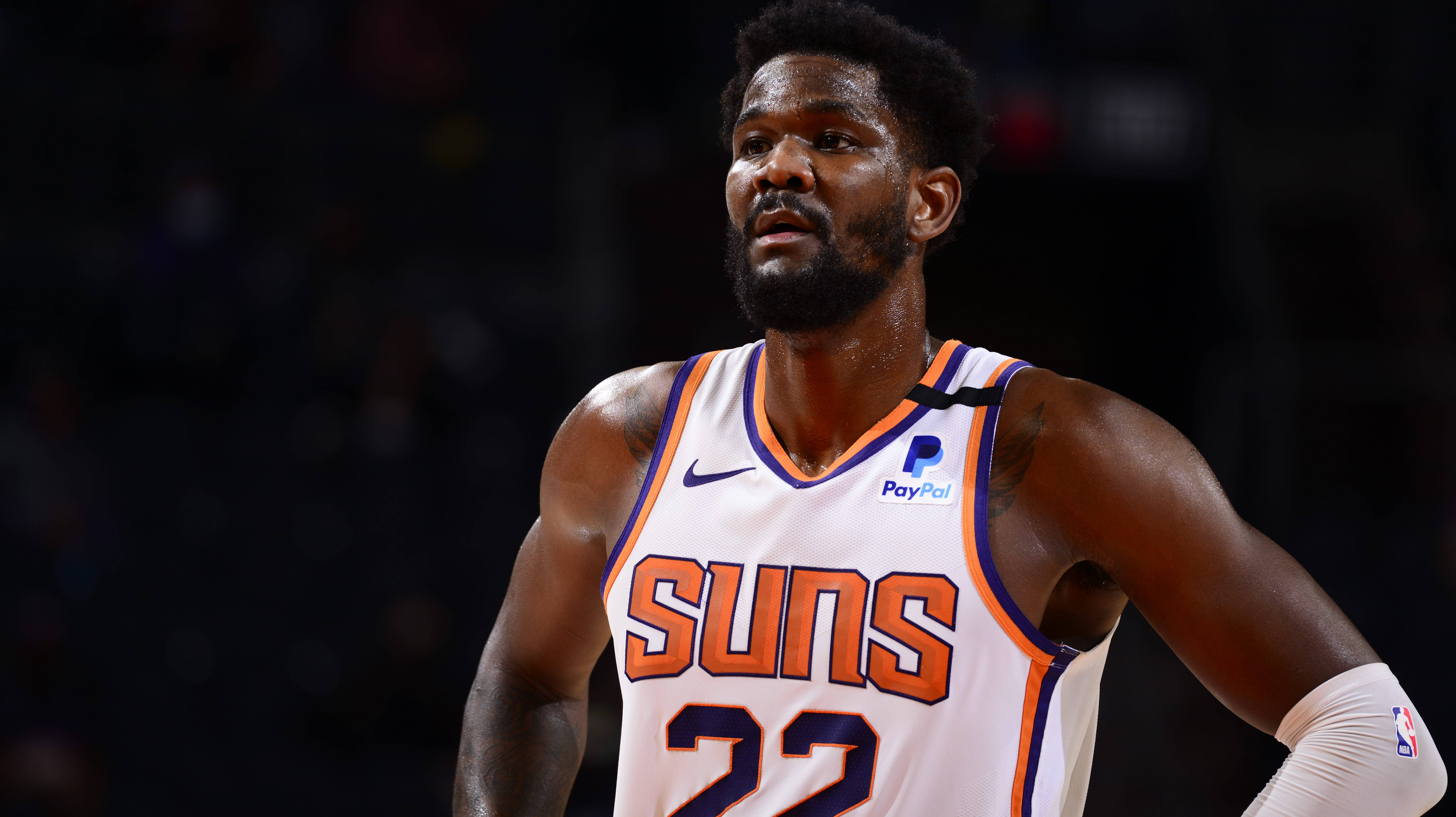 NBA 桌布 壁紙 — 一名專注的籃球運動員身穿白色Suns球衣，帶有橙色和紫色飾邊，在比賽期間站在燈光昏暗的球場上。