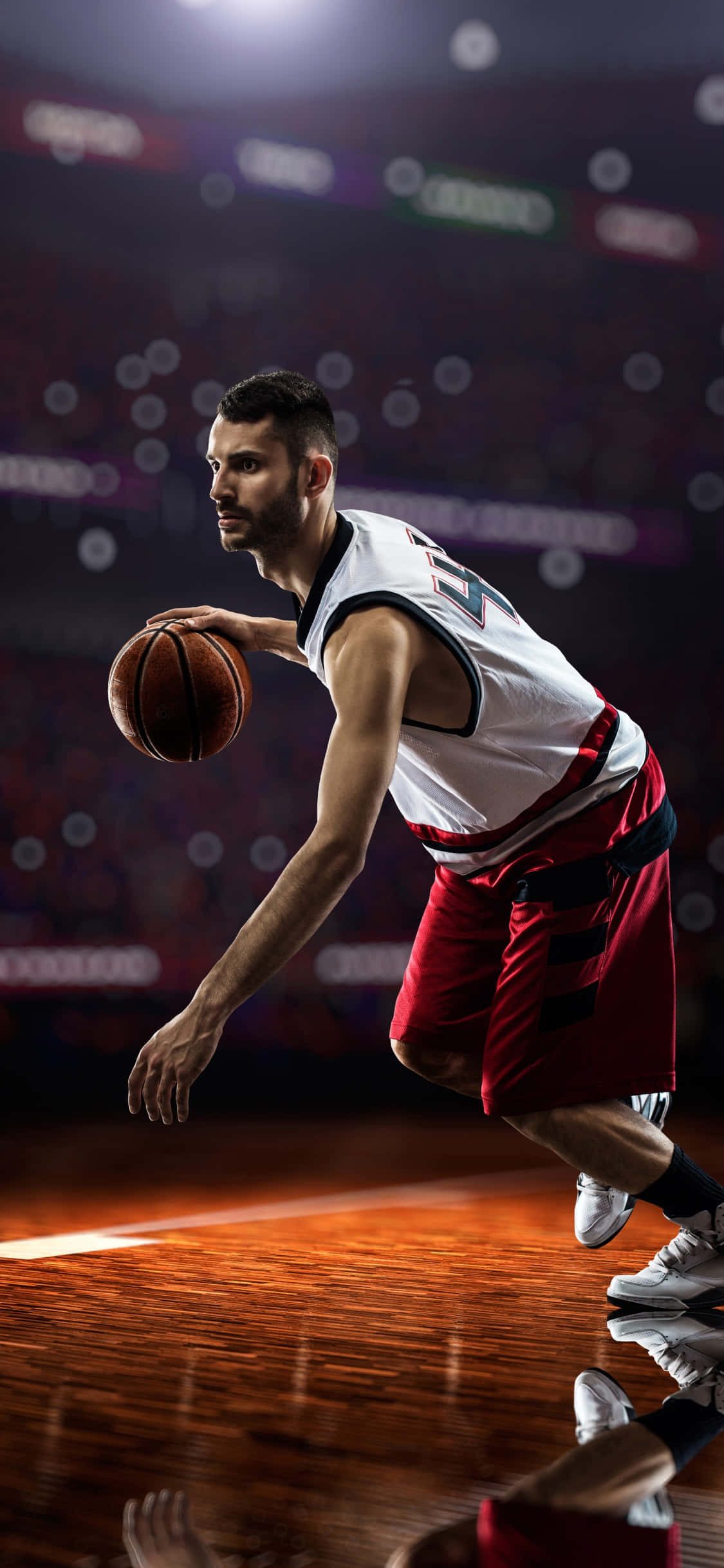 iPhone Xs Basketball 桌布 壁紙 — 一名專注的籃球運動員身穿白色和紅色制服，在明亮的場館燈光下激烈運球，背景是模糊的人群。