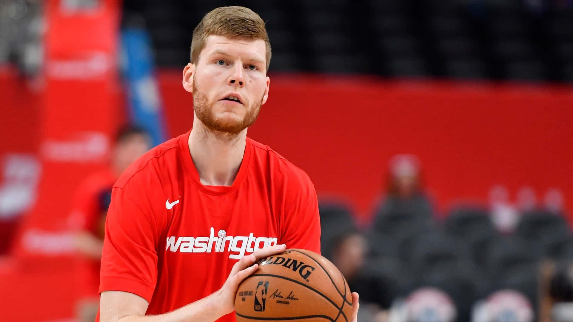 NBA 桌布 壁紙 — 一名專注的籃球運動員身穿紅色華盛頓球衣，在比賽中手持籃球，背景是模糊的體育館座位。