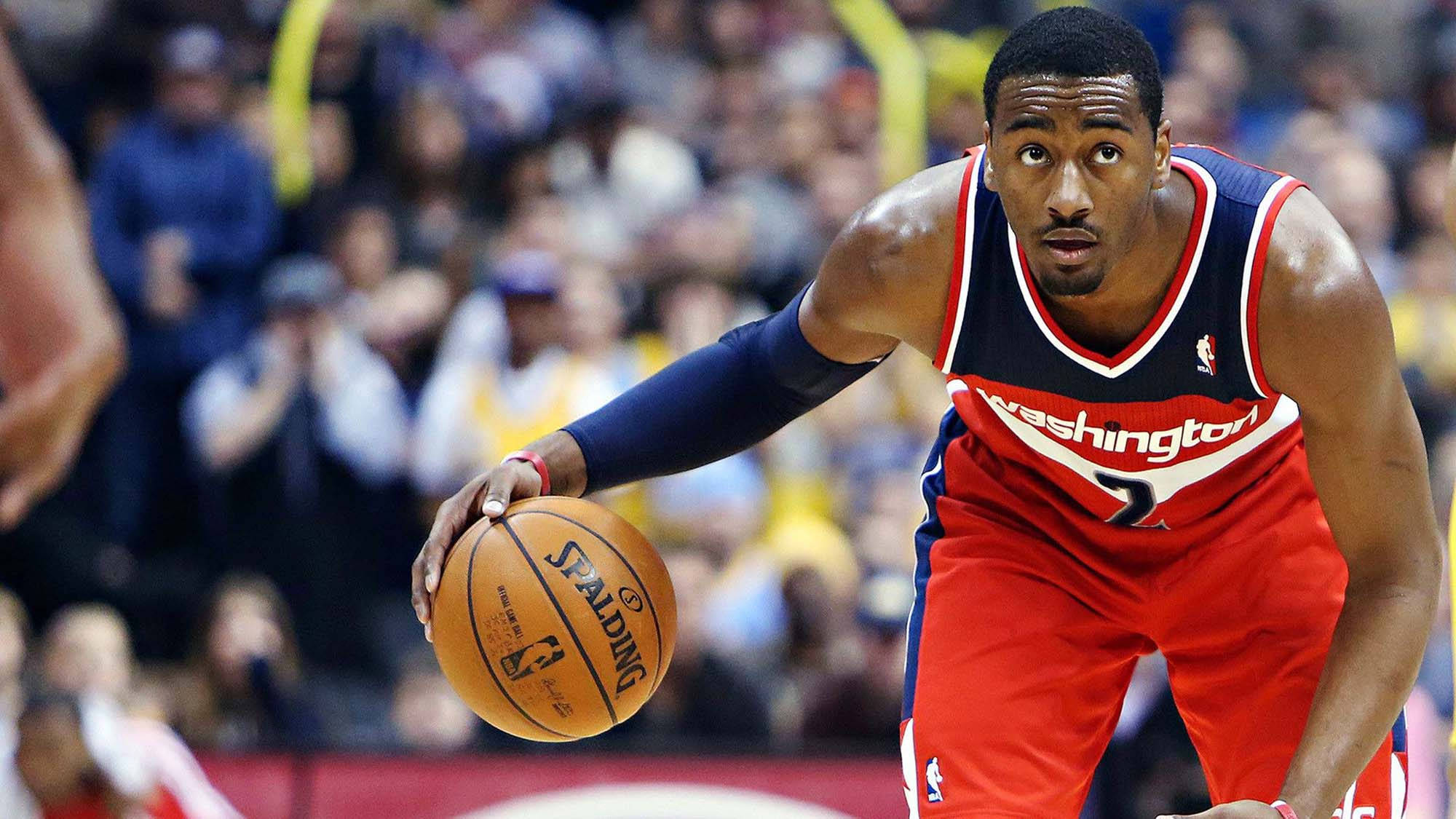 John Wall 桌布 壁紙 — 一名專注的籃球運動員身穿紅色華盛頓球衣，在激烈的比賽中運著Spalding籃球，身後是模糊的人群。