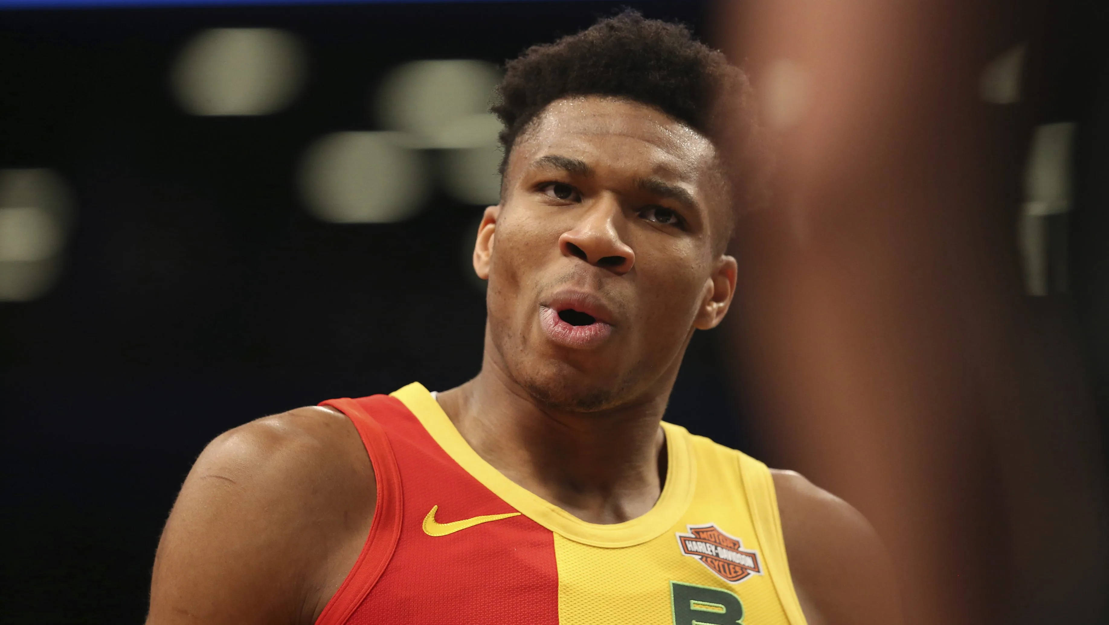 Giannis 桌布 壁紙 — 一名專注的籃球運動員身穿紅黃球衣，在明亮的場館燈光下比賽中凝視著。