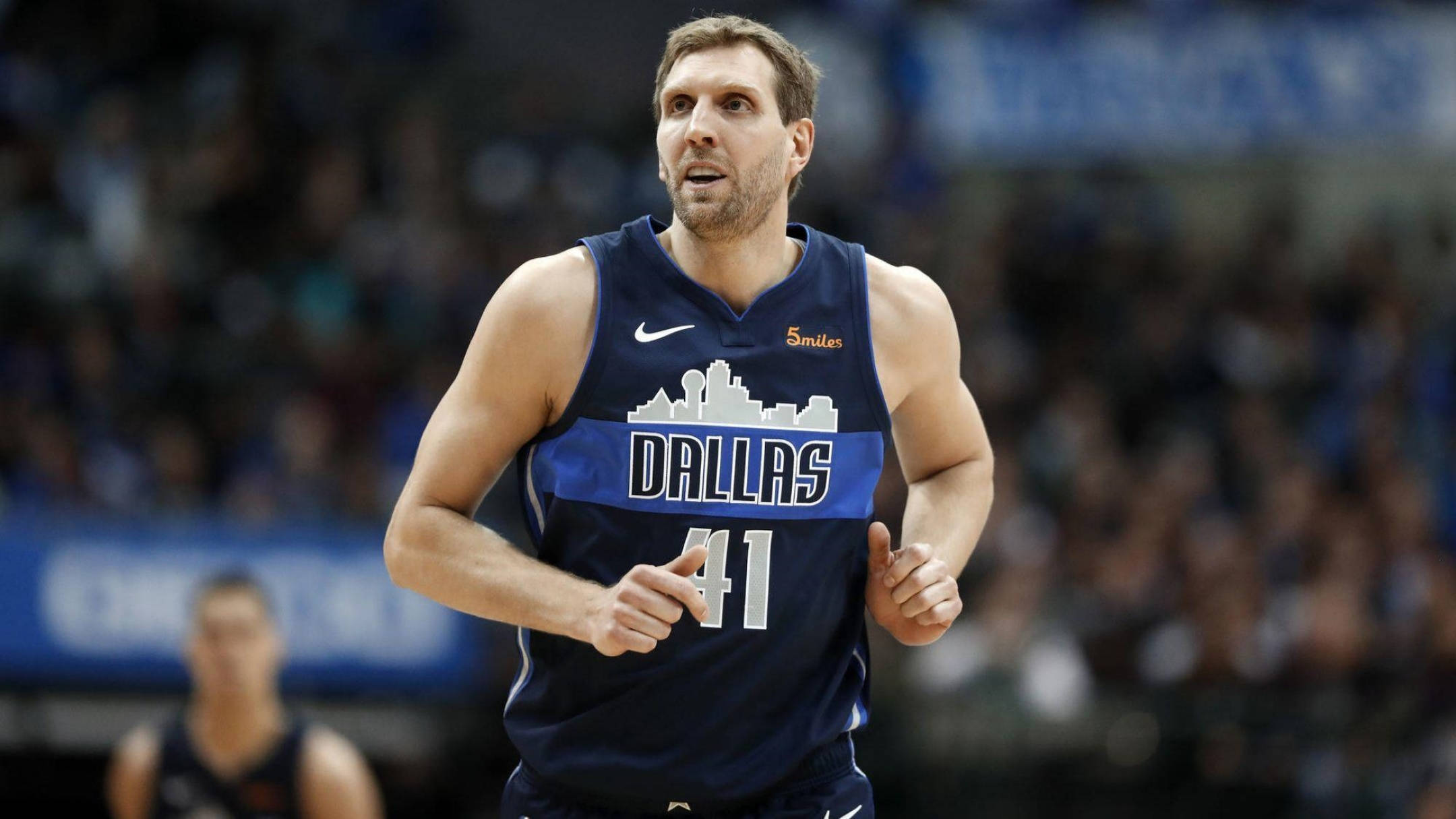 Dirk Nowitzki 桌布 壁紙 — 一名專注的籃球運動員身穿達拉斯獨行俠球衣，在比賽中衝刺，背景是模糊的觀眾。