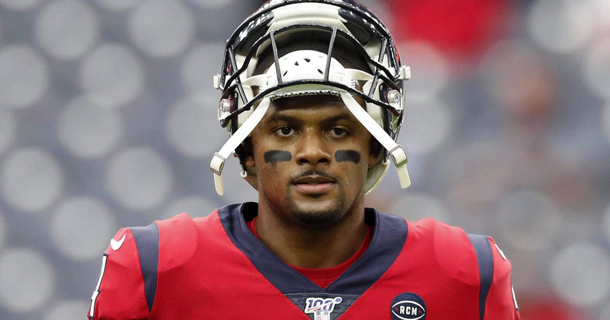 Deshaun Watson 桌布 壁紙 — 一名專注的美式足球員身穿紅色球衣和頭盔，帶著黑色眼漆，在模糊的體育場背景下凝視著。