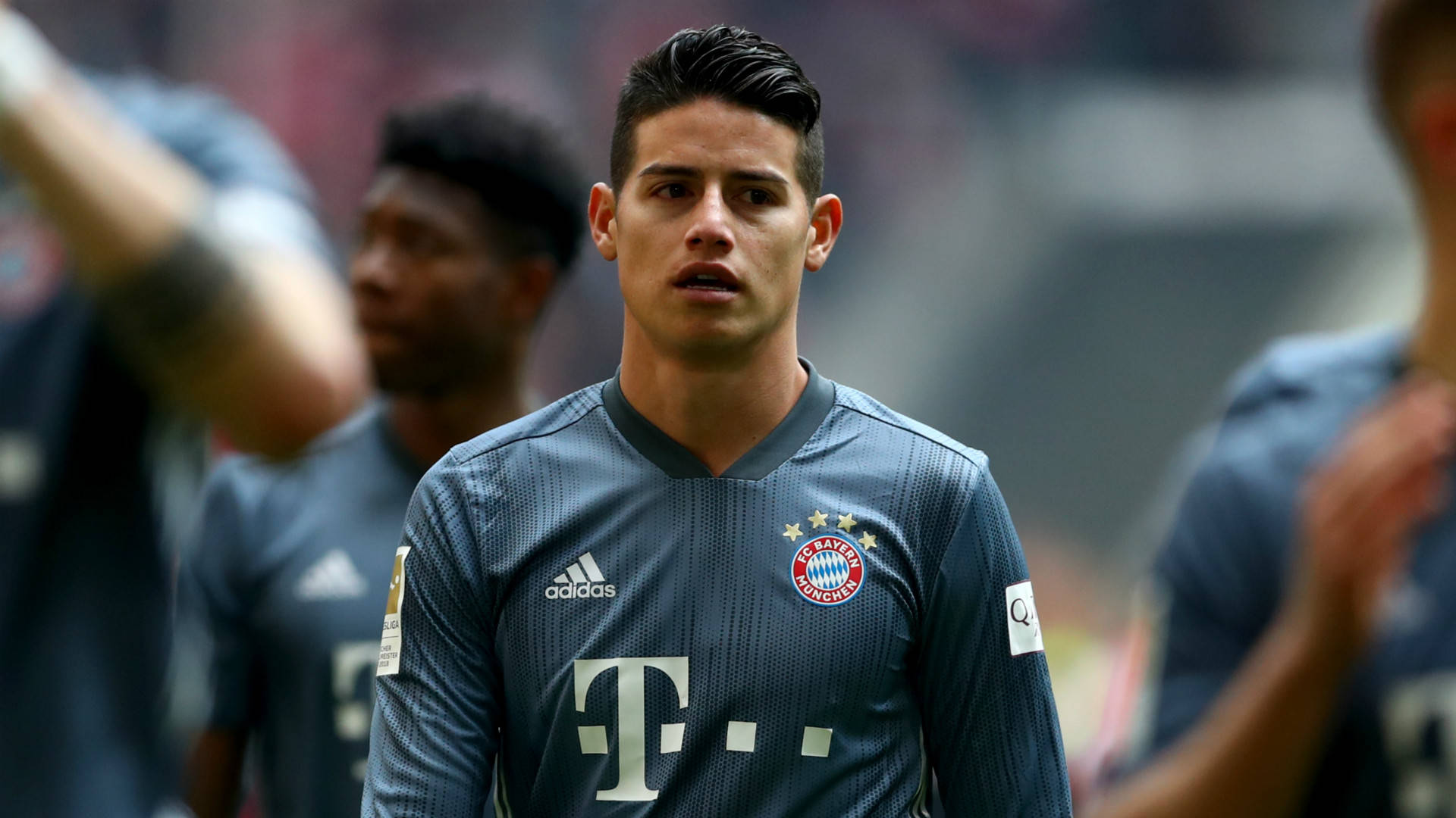 James Rodriguez 桌布 壁紙 — 一名專注的足球員身穿深藍色拜仁慕尼黑球衣，站在陽光明媚的球場上，背景中的隊友模糊不清。