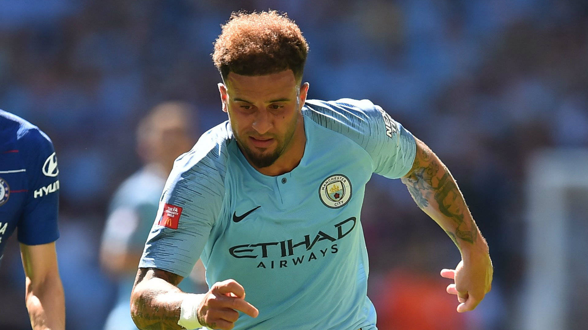 Kyle Walker 桌布 壁紙 — 一名專注的足球員身穿淺藍色曼城球衣，在日間比賽中衝刺，身後是模糊的球場人群。