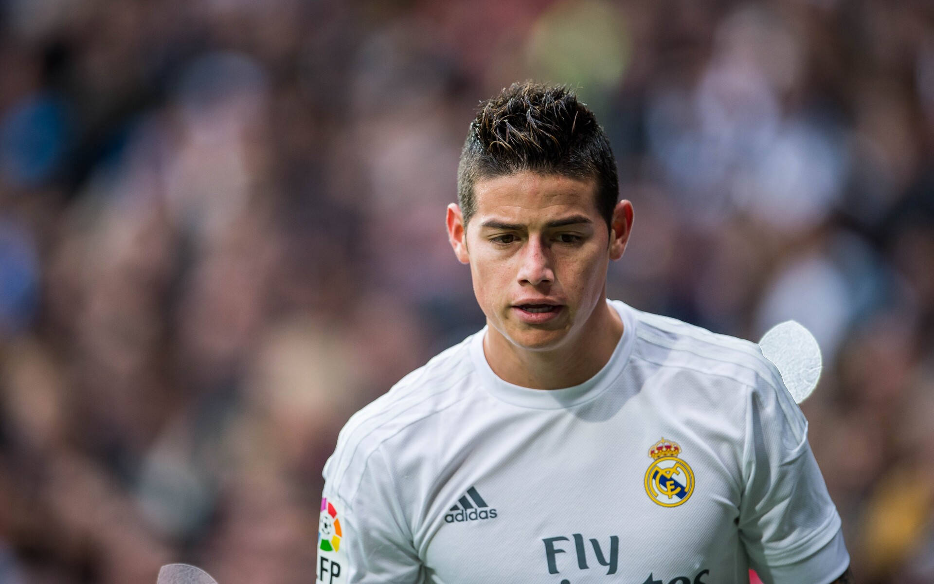 James Rodriguez 桌布 壁紙 — 一名專注的足球員身穿白色皇家馬德里球衣，站在陽光普照的球場上，背後是模糊的體育場人群。