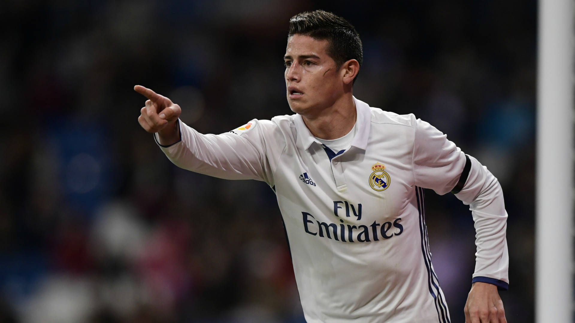 James Rodriguez 桌布 壁紙 — 一名專注的足球員身穿白色皇家馬德里球衣，在球場燈光下的夜間比賽中果斷地指向某處。