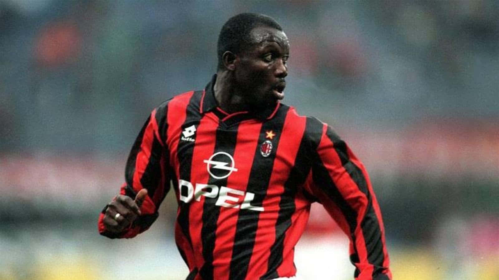 Ac Milan 桌布 壁紙 — 一名專注的足球員身穿紅黑條紋球衣，在模糊的球場上衝刺。