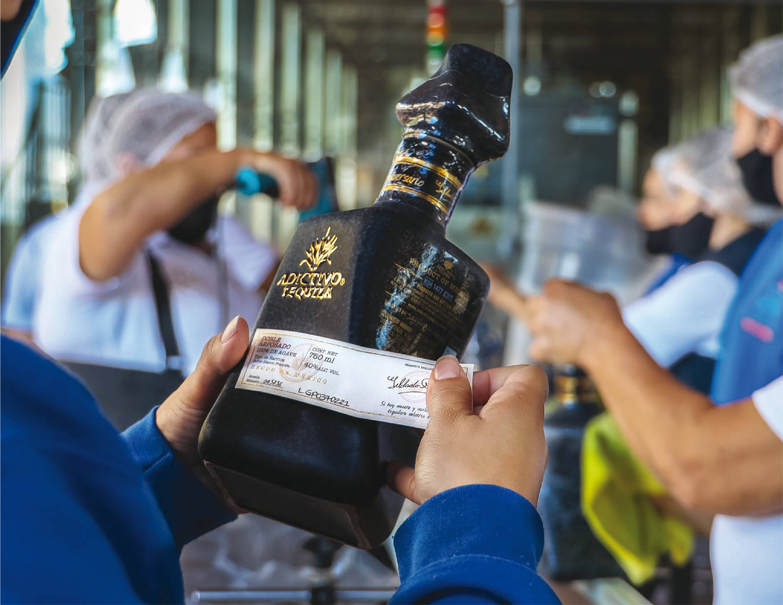 筆電 桌布 壁紙 — 一名工人手持一瓶帶有金色字樣的黑色Adictivo Tequila，背景中工廠員工正在工作。