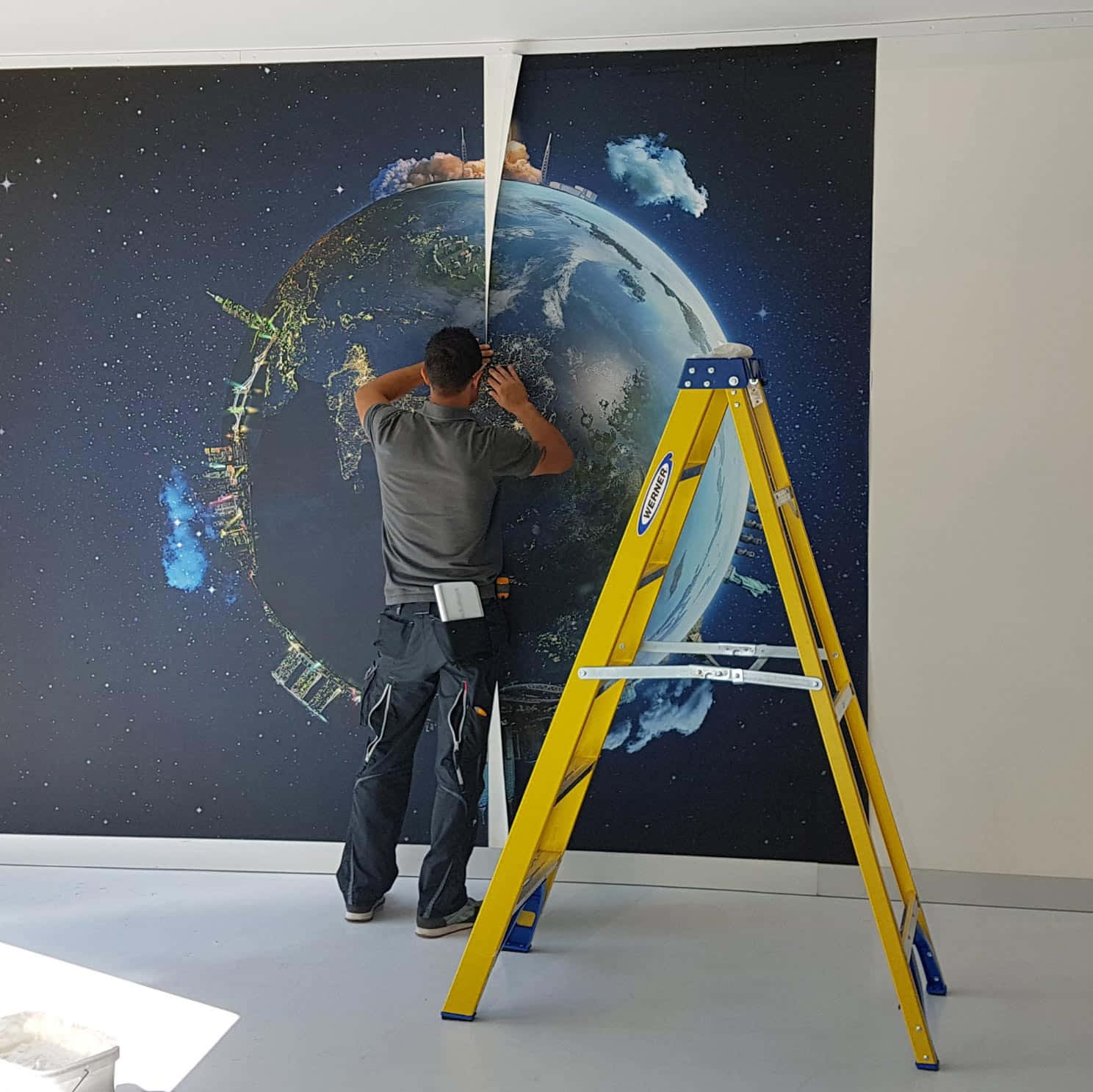 Installation Art 桌布 壁紙 — 一名工人正在安裝一幅大型太空主題壁紙，上面有地球和星星，旁邊有一把黃色梯子，牆壁是素色的。