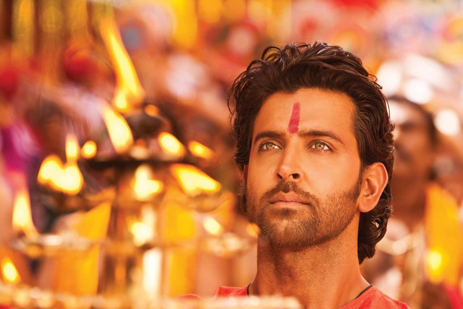 Bollywood Hero 桌布 壁紙 — 一名帶有紅色眉心貼的男子在充滿活力的寺廟環境中，在閃爍的金色迪亞燈中向上凝視。