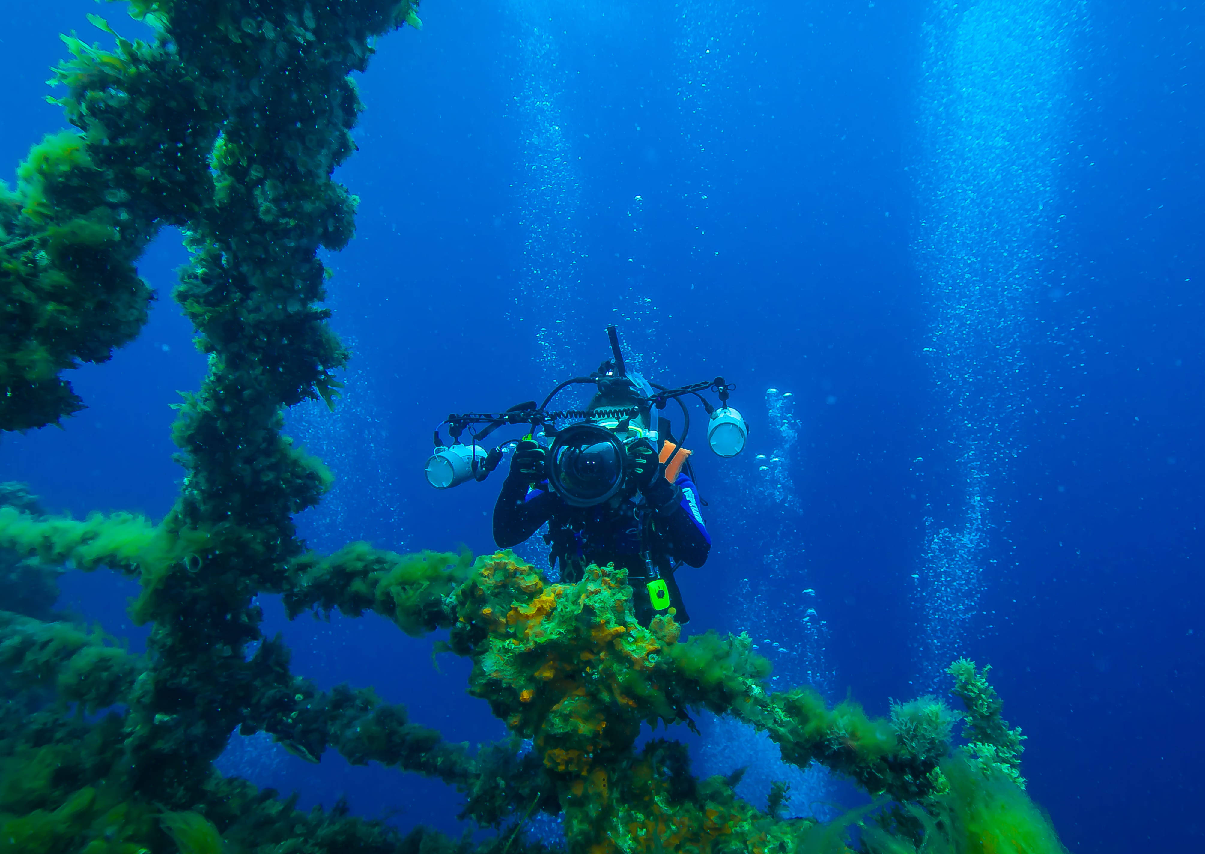 diving 桌布 壁紙 — 一名帶著大型相機的水肺潛水員在深藍色海水中探索一個陽光普照、被珊瑚覆蓋的沉船。