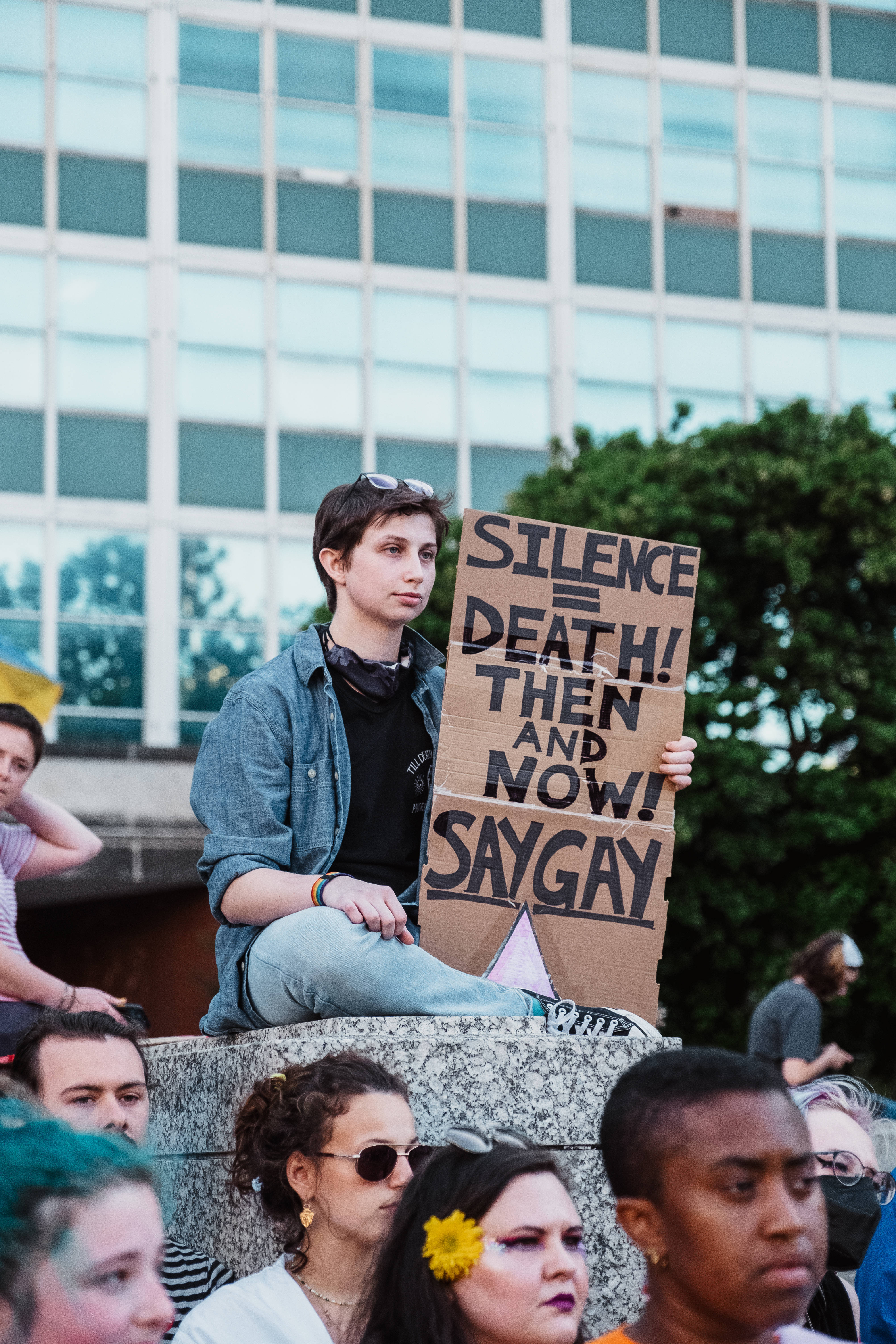 Binary 桌布 壁紙 — 一名年輕人坐在石階上，在戶外集會中舉著寫有「silence equals death then and now saygay」的抗議標語牌。