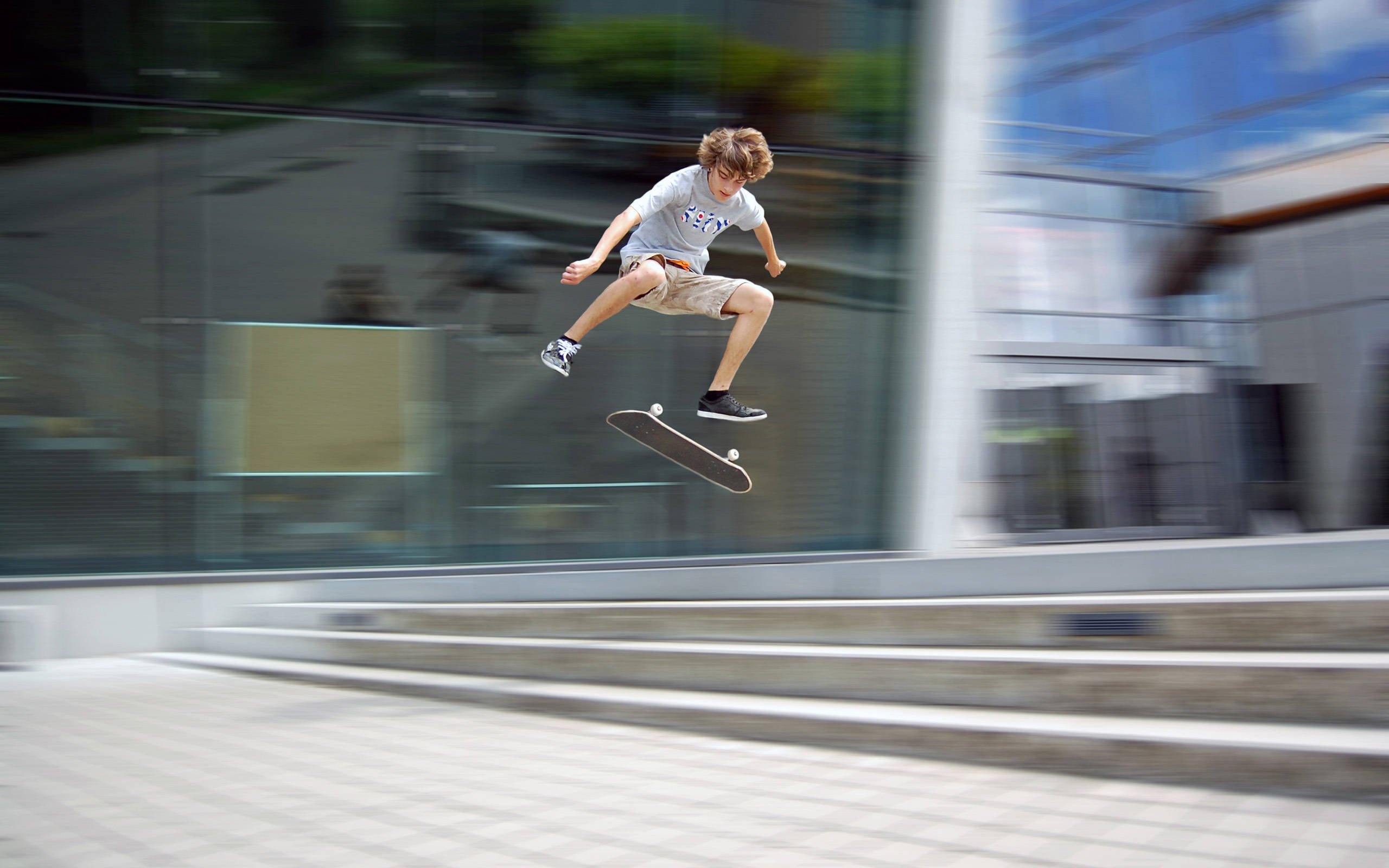 Skater Boy 桌布 壁紙 — 一名年輕滑板手在半空中表演花式，背景是模糊的現代玻璃建築。