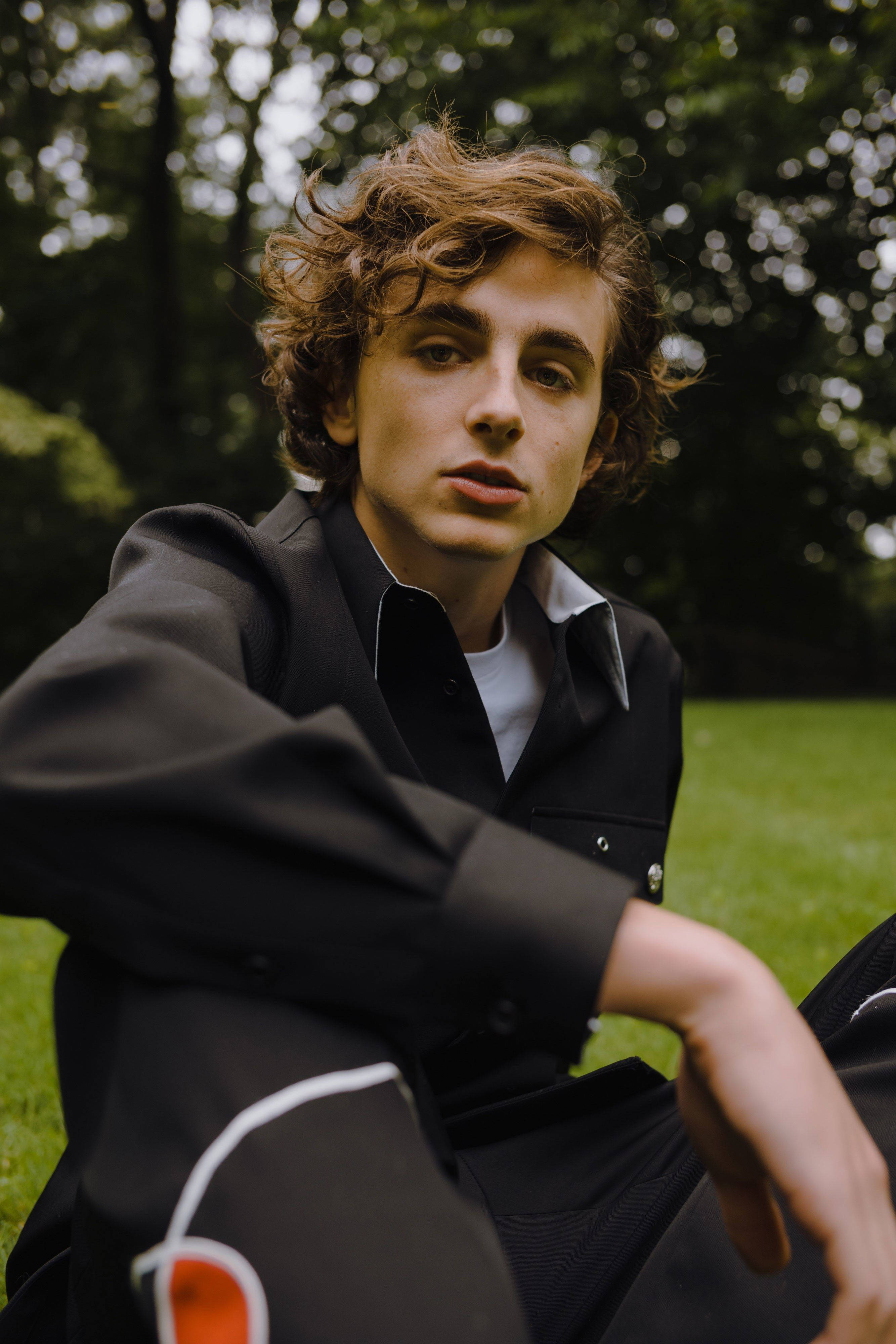 Timothée Chalamet 桌布 壁紙 — 一名年輕男子，棕色捲髮，坐在森林的草地上，身穿黑色外套內搭白色襯衫，直視鏡頭。