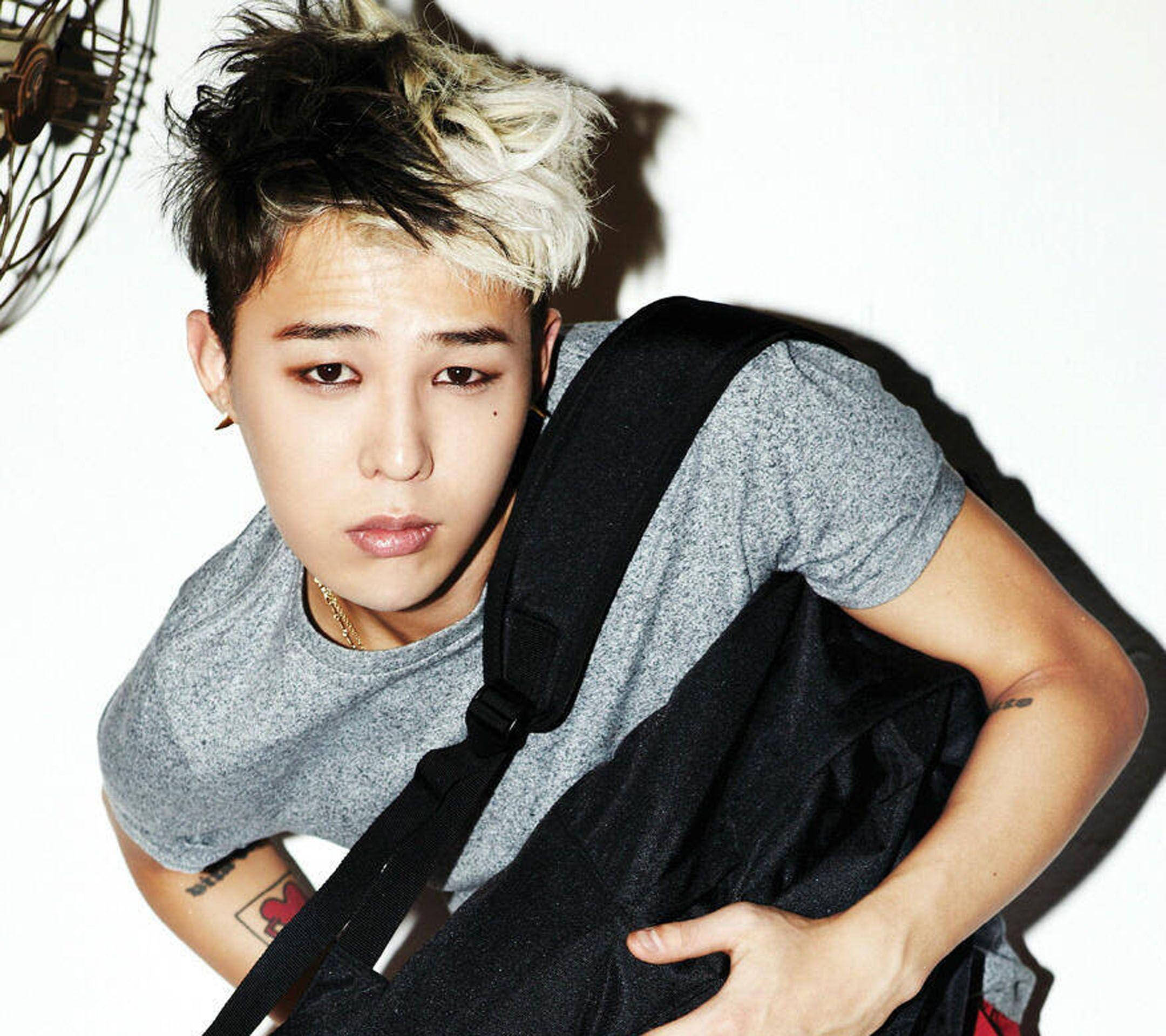 G-dragon 桌布 壁紙 — 一名年輕男子，頭髮黑白尖刺，身穿灰色T恤和黑色背包，在純白背景前向前傾。