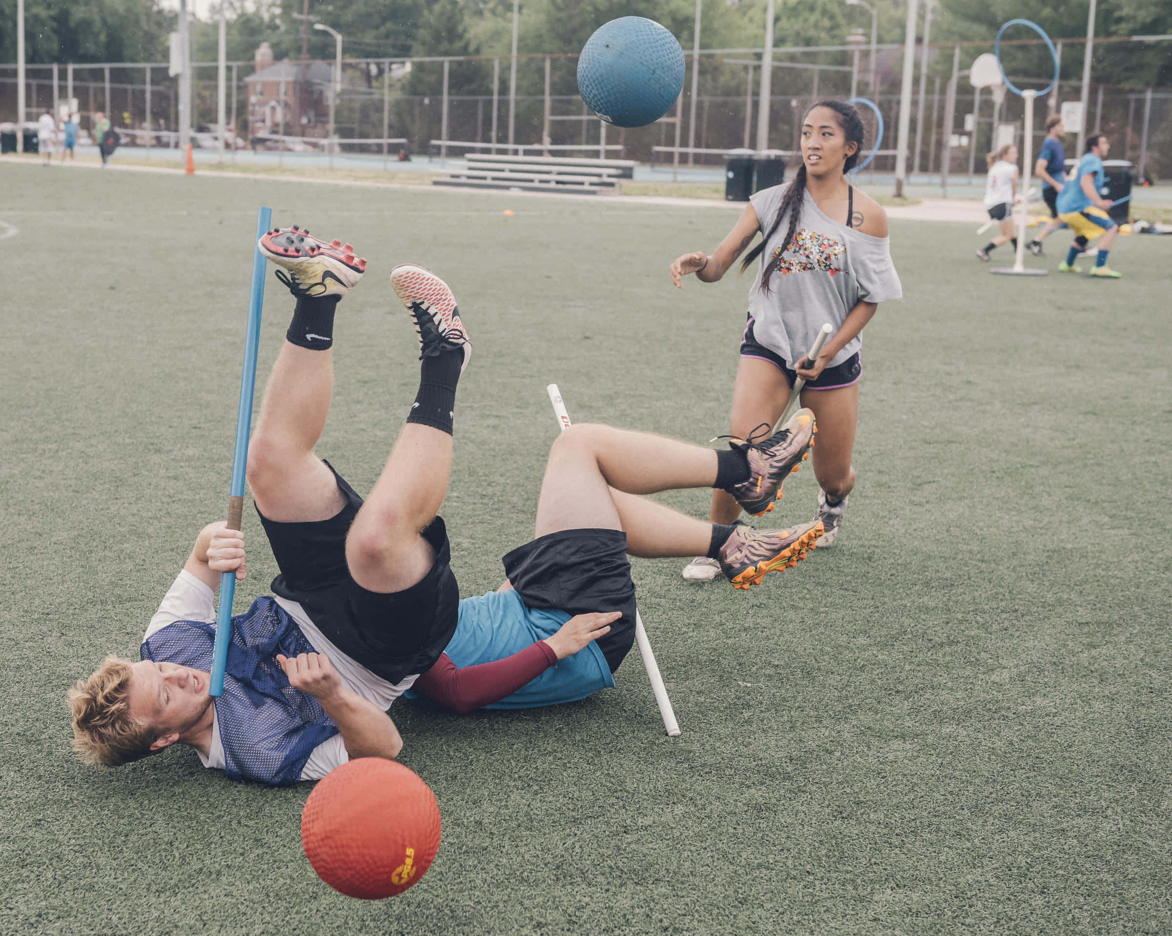 Quidditch 桌布 壁紙 — 一名年輕男子仰臥在運動場上踢一個藍色球，而一名女子則拿著長曲棍球棍觀看。