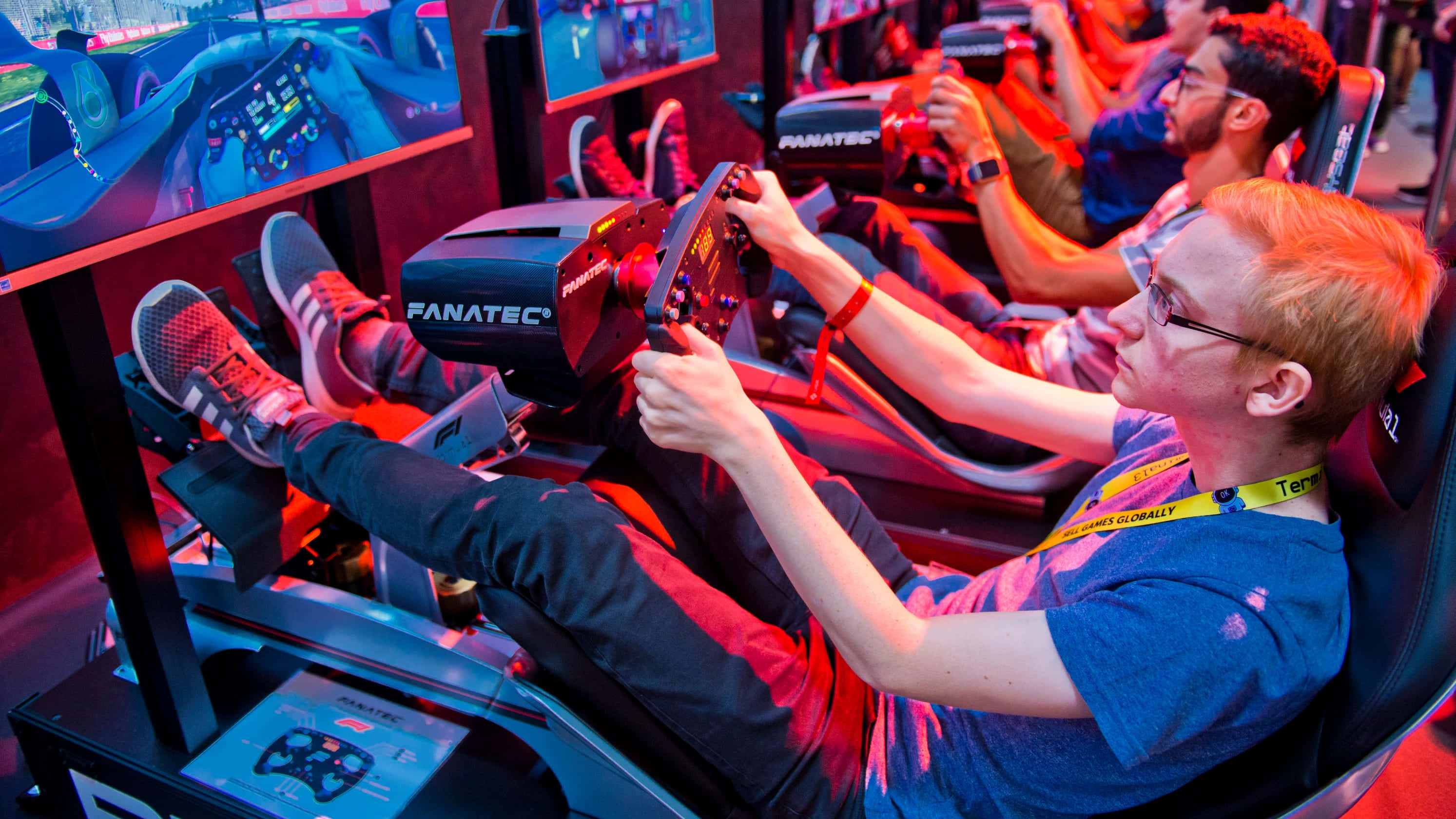 Gaming Conventions 桌布 壁紙 — 一名年輕男子在Fanatec賽車模擬器上專注地玩賽車遊戲，紅色燈光閃爍，背景有其他玩家。