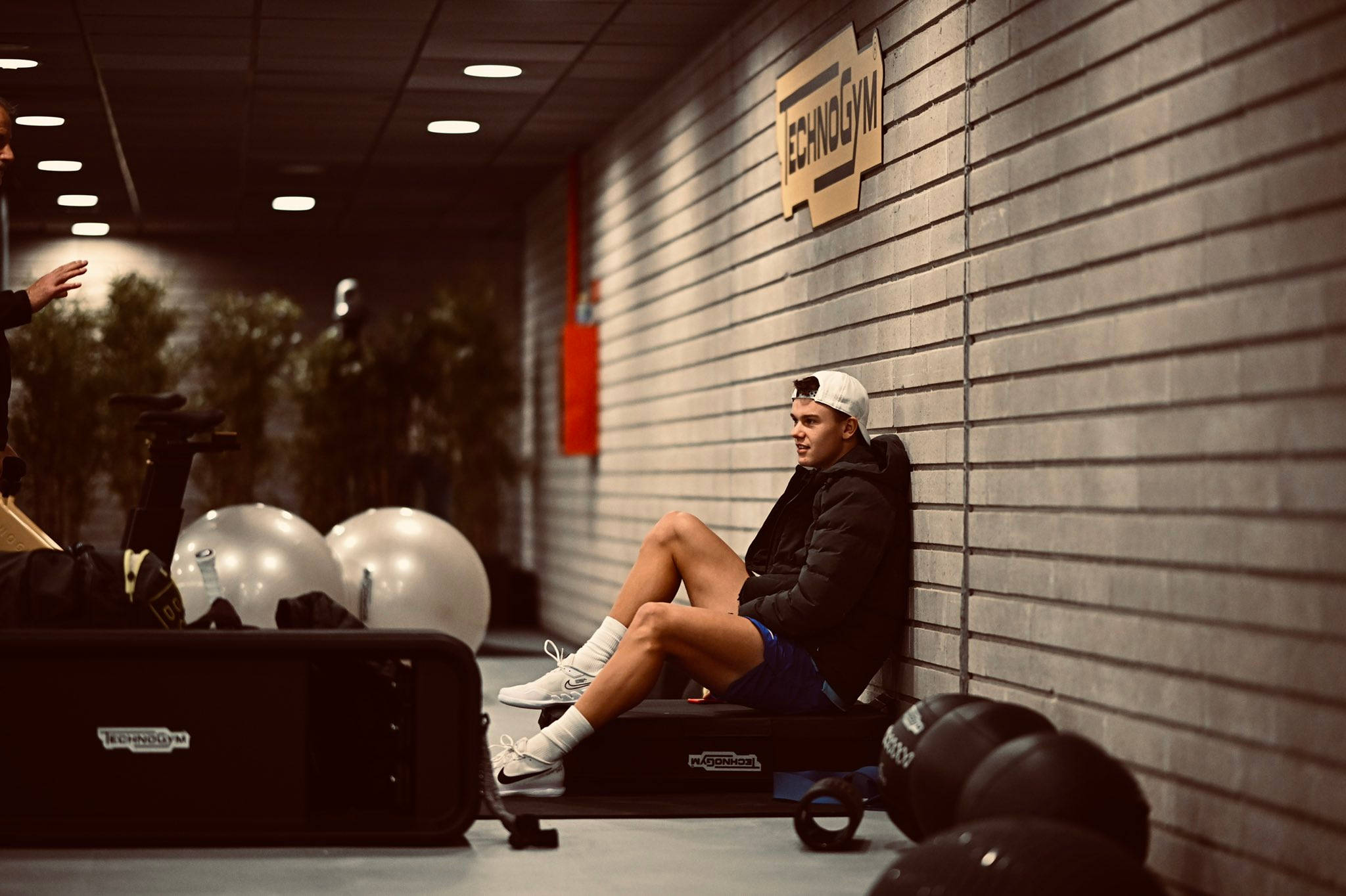 Champion 桌布 壁紙 — 一名年輕男子在健身房內靠著磚牆坐在長凳上休息，而教練在附近做手勢。