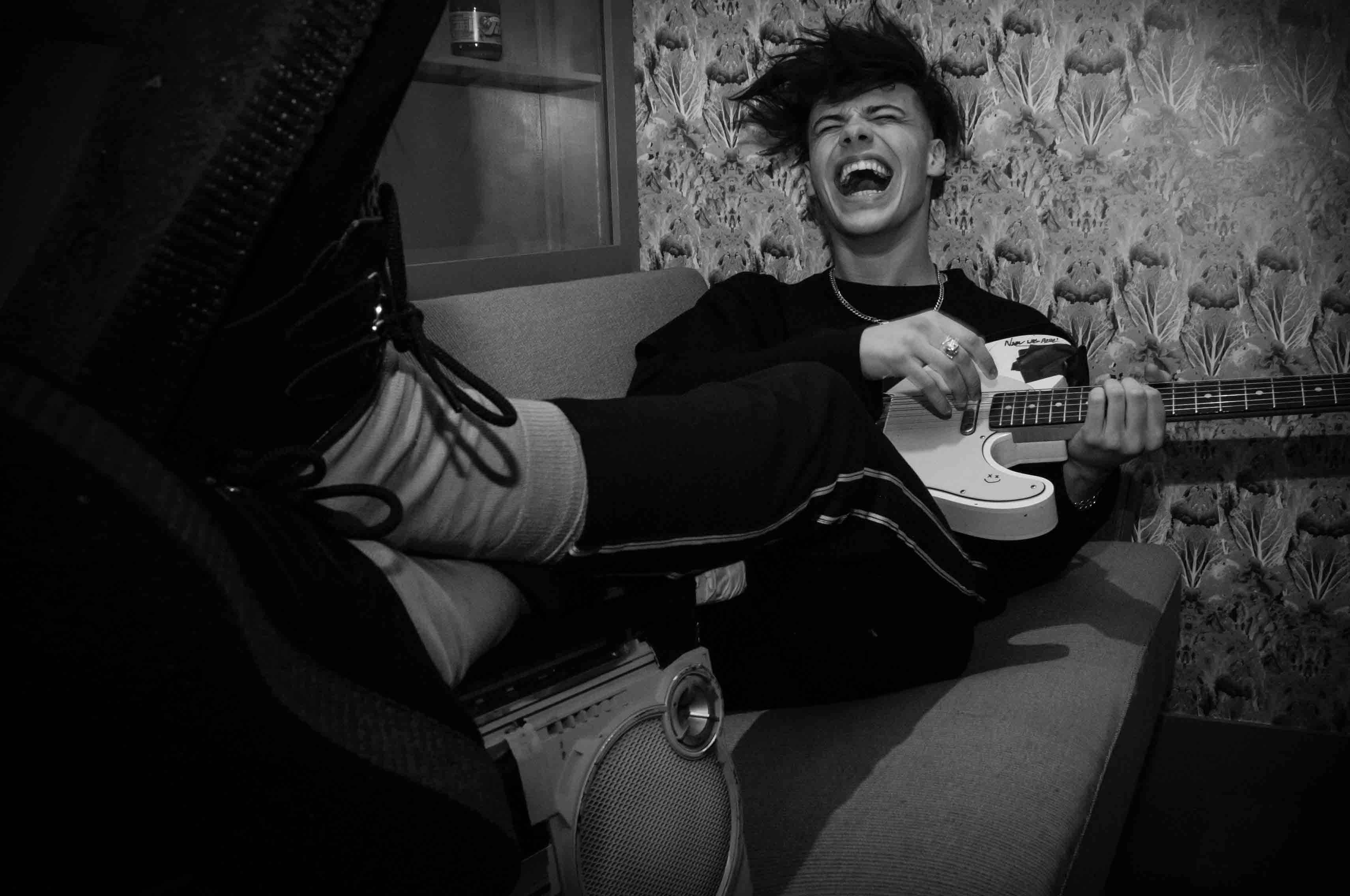Yungblud 桌布 壁紙 — 一名年輕男子在圖案壁紙前的沙發上彈奏白色電吉他，狂放大笑。