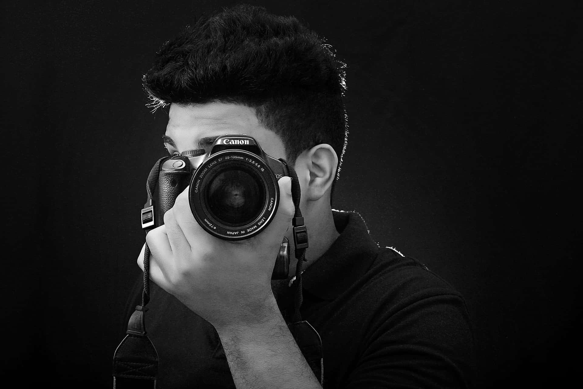 Dslr 桌布 壁紙 — 一名年輕男子在強烈的黑色背景下，將佳能數位單眼相機舉到眼前，拍攝出一幅戲劇性的黑白肖像。
