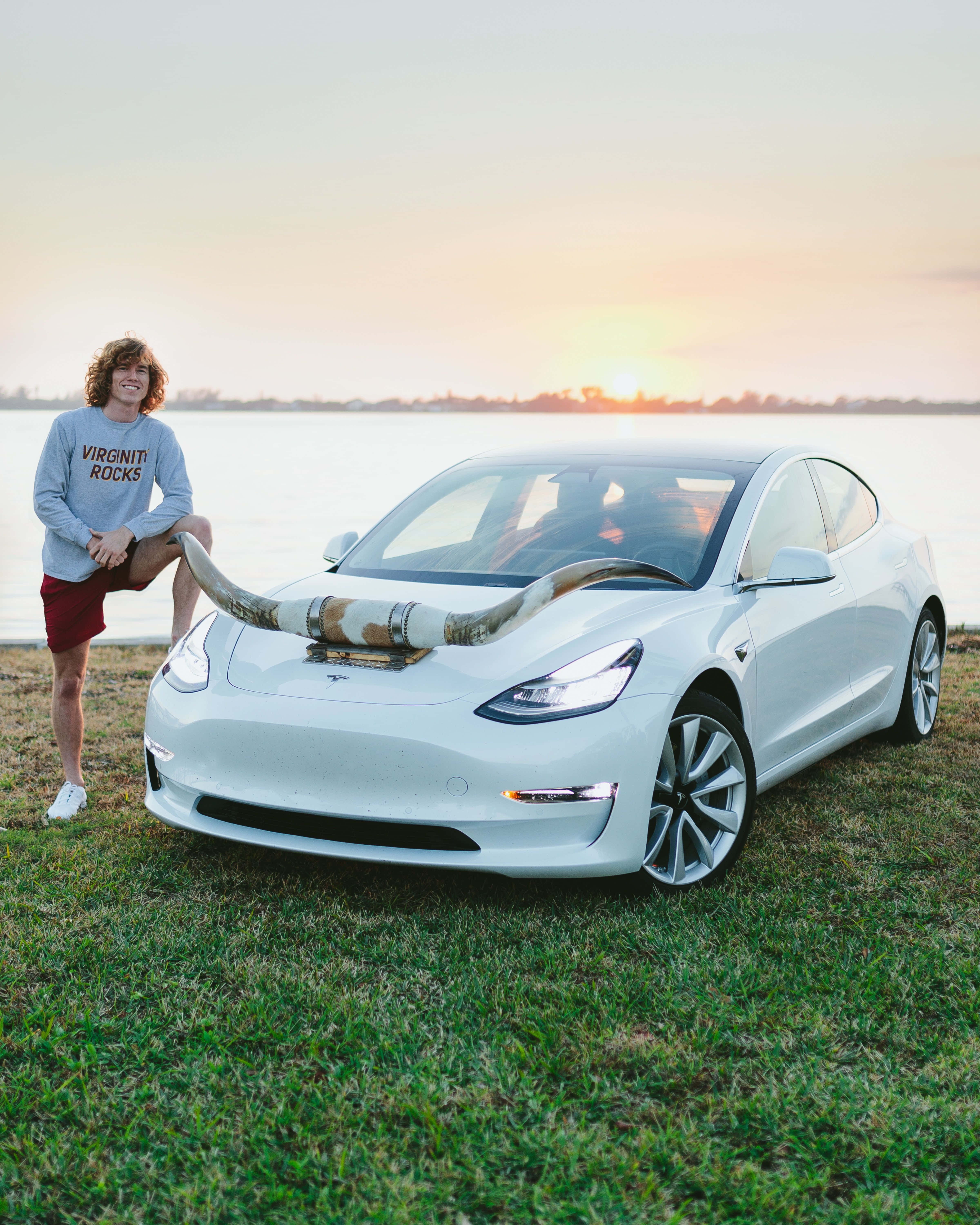 日落 桌布 壁紙 — 一名年輕男子在白色Tesla Model 3旁微笑，車上裝有公牛角，停泊在日落時分近水的草地上。