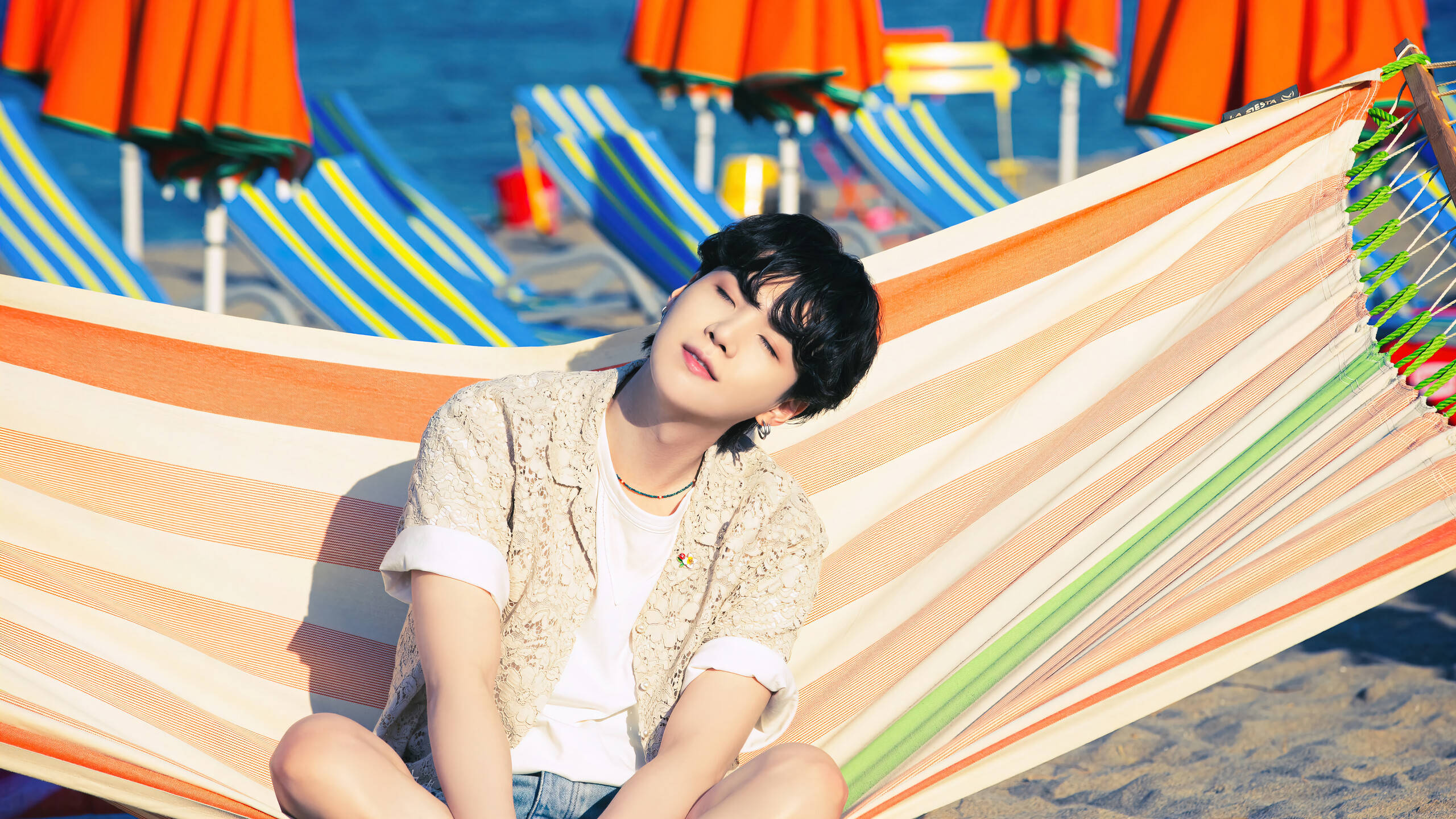 Suga 桌布 壁紙 — 一名年輕男子在陽光明媚的海灘上，背景有彩色遮陽傘和躺椅，他正平靜地躺在條紋吊床上。