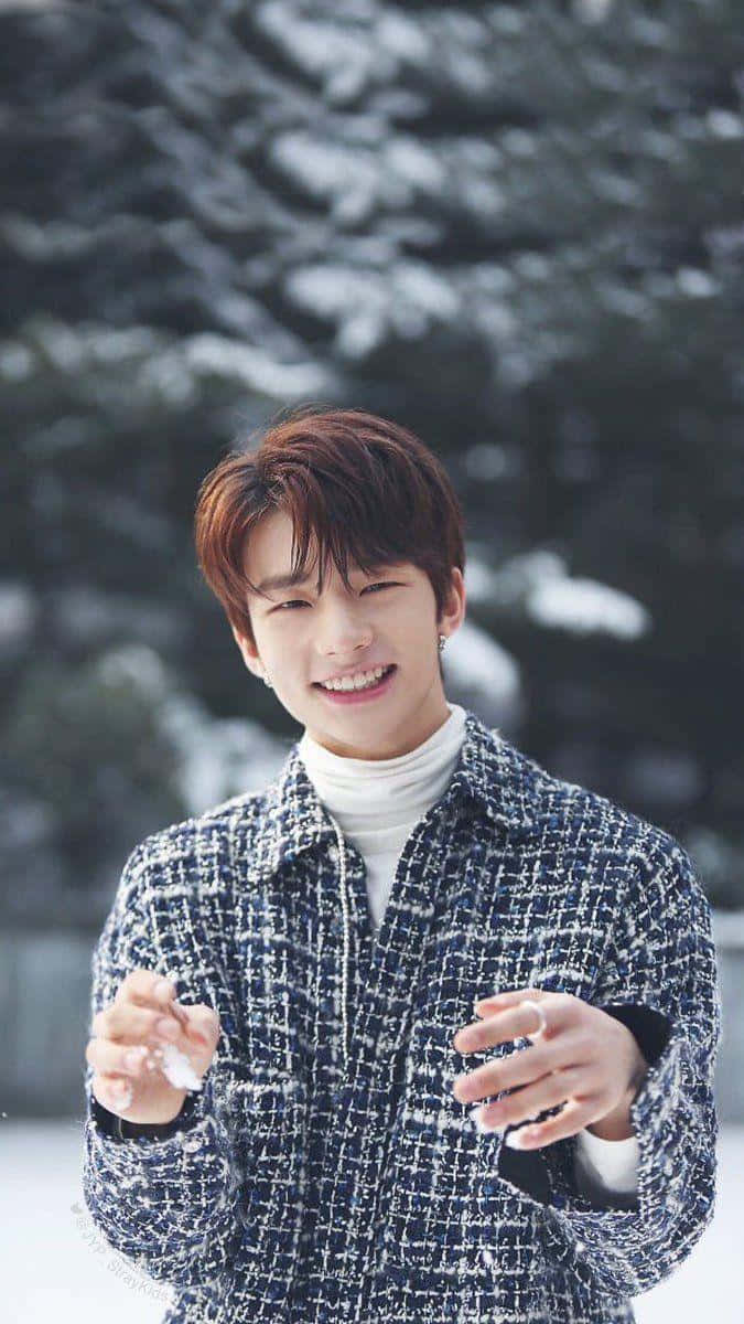 hyunjin 桌布 壁紙 — 一名年輕男子在雪地森林背景下，雙手捧著雪花，露出燦爛的笑容。