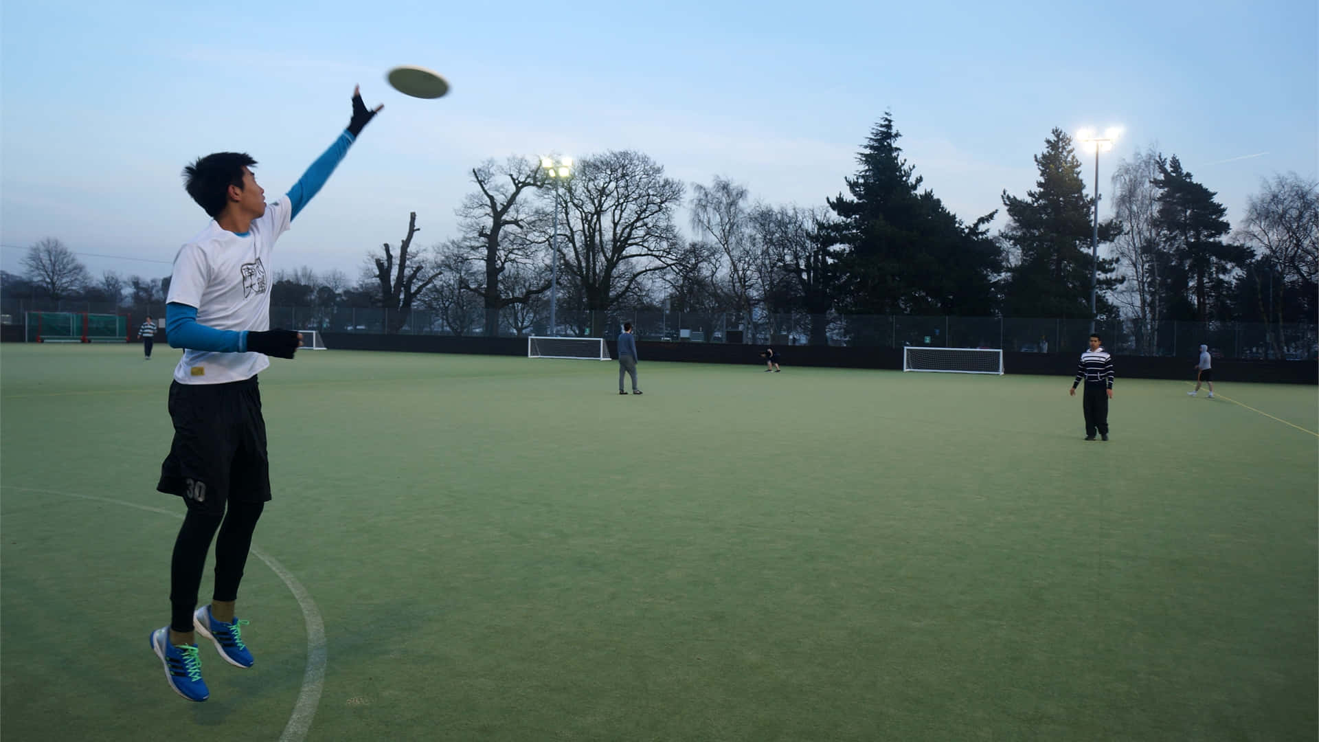 Frisbee 桌布 壁紙 — 一名年輕男子在黃昏時分的綠色運動場上跳起接飛盤，背景有光禿的樹木和體育場燈光閃爍。