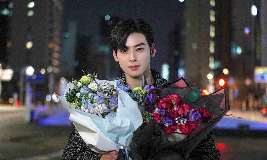 True Beauty 桌布 壁紙 — 一名年輕男子夜晚站在城市中，手持兩大束色彩繽紛的花朵，背景是模糊的城市景觀。