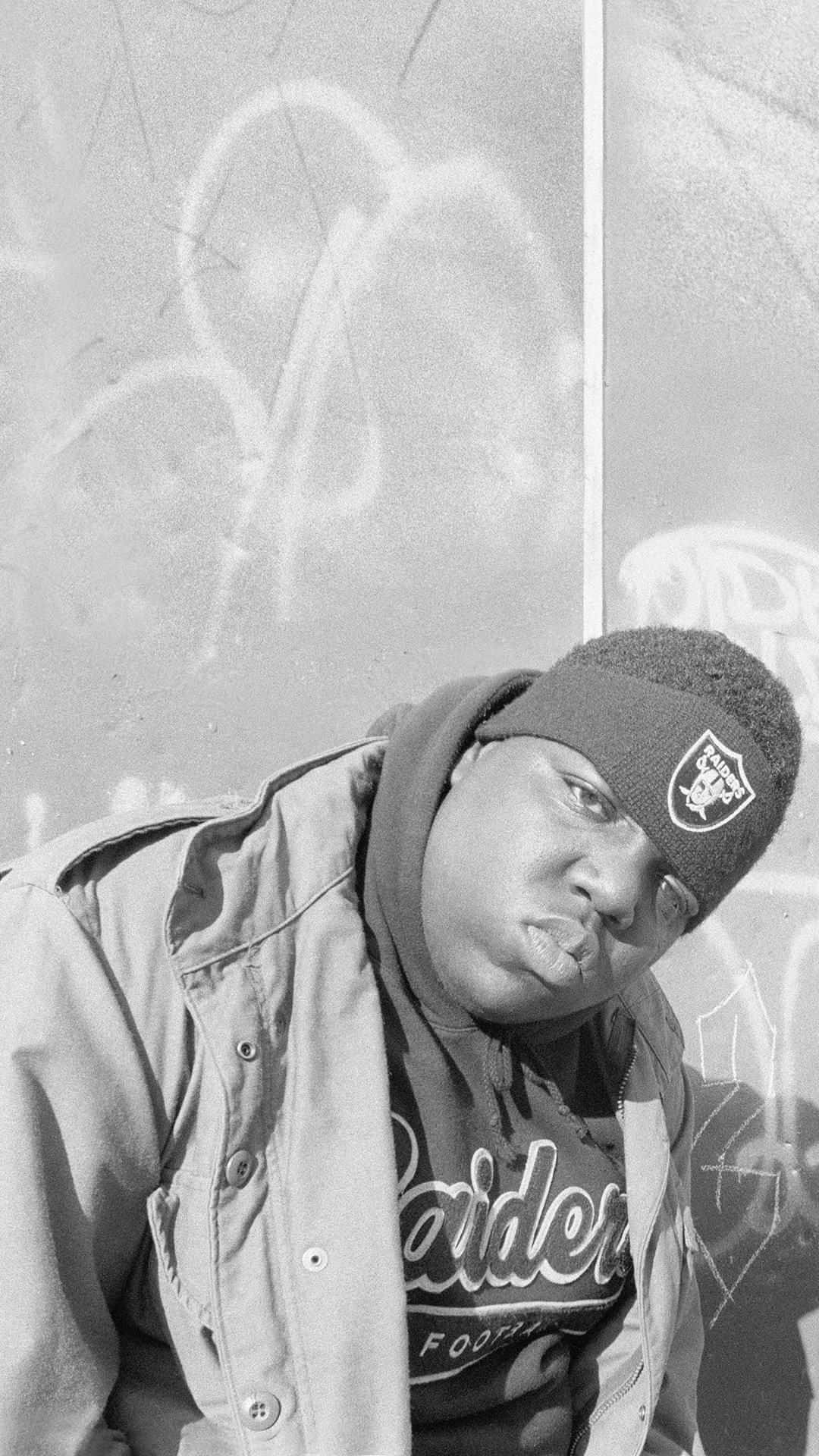 Rappers 桌布 壁紙 — 一名年輕男子戴著Raiders毛帽和穿著外套，靠在一面塗鴉牆上，呈現粗糙的黑白風格。