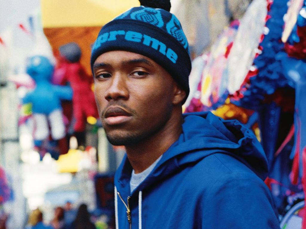 Frank Ocean 桌布 壁紙 — 一名年輕男子戴著藍色Supreme毛帽和連帽衫，在充滿活力、模糊的街市背景下凝視著鏡頭。