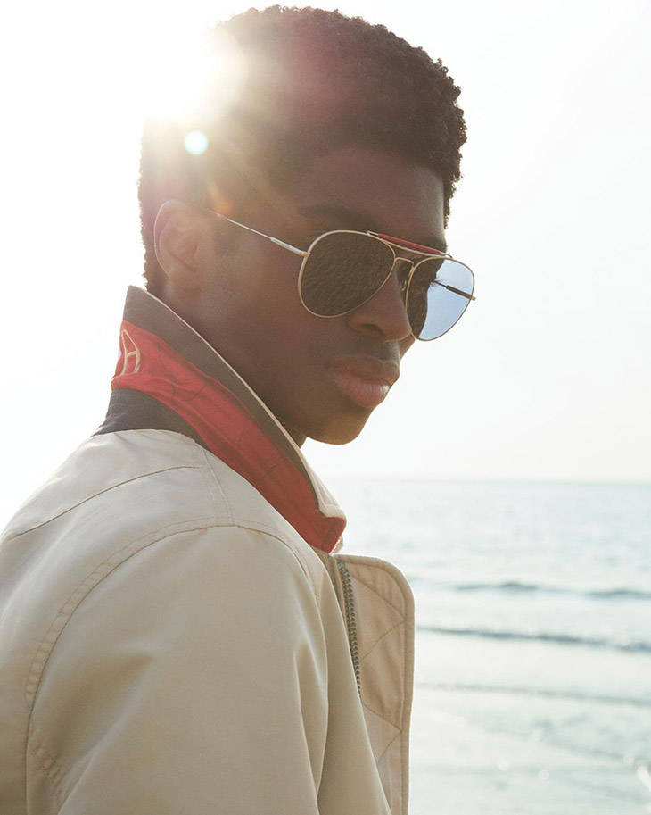 Sunglasses 桌布 壁紙 — 一名年輕男子戴著飛行員太陽眼鏡和米色外套站在海邊，明亮的陽光在他頭後形成眩光。