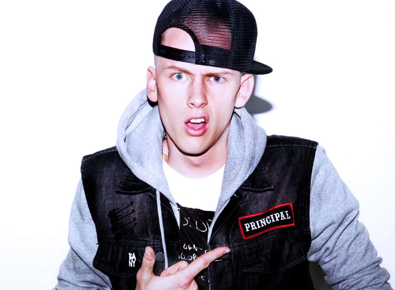 mgk 桌布 壁紙 — 一名年輕男子戴著黑色網帽和牛仔背心，指著一個「PRINCIPAL」補丁，在白色背景前露出驚訝的表情。