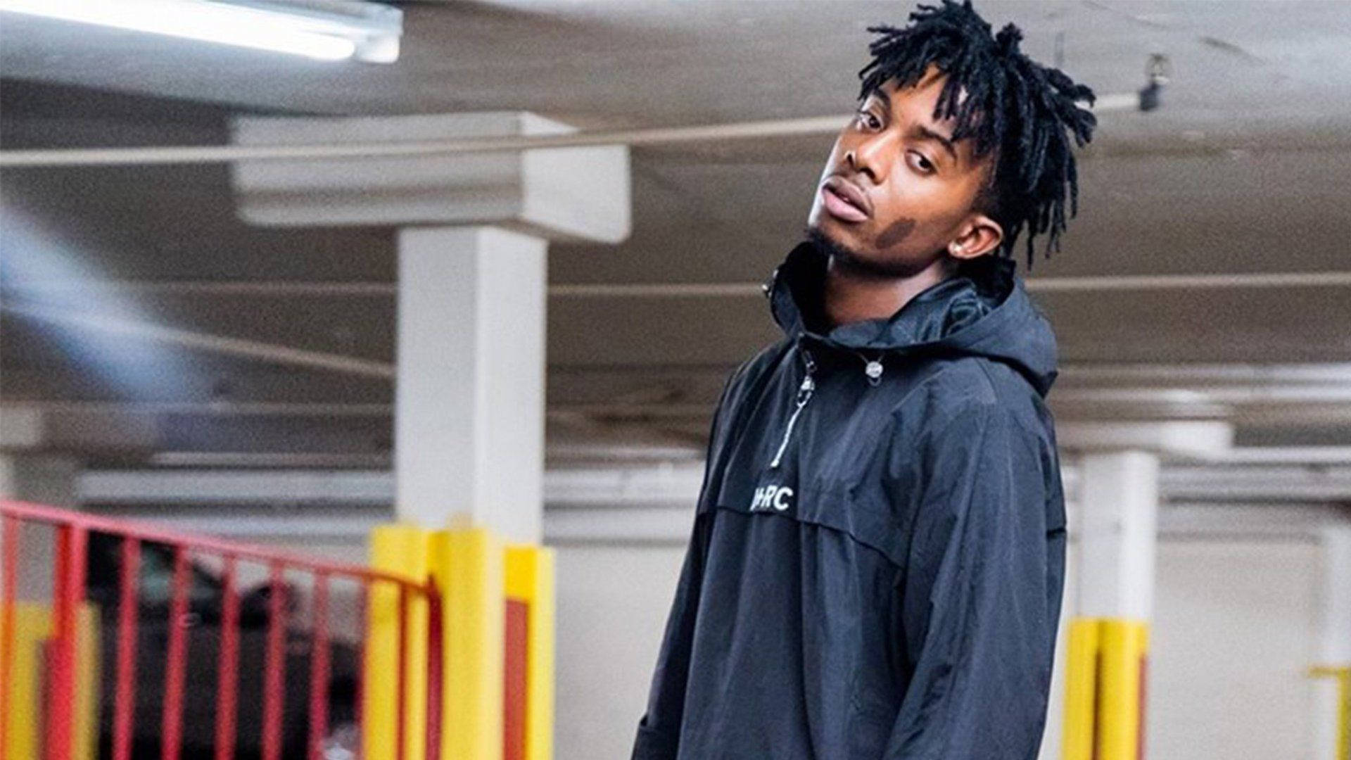 Carti 桌布 壁紙 — 一名年輕男子梳著辮子頭，身穿黑色外套，站在光線昏暗的停車場內，周圍有紅色和黃色的欄杆。