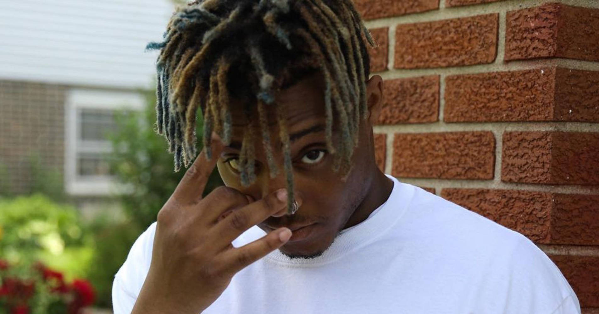 年輕男子留著色彩繽紛的辮子，在磚牆附近做出和平手勢。 — Juice Wrld 壁紙