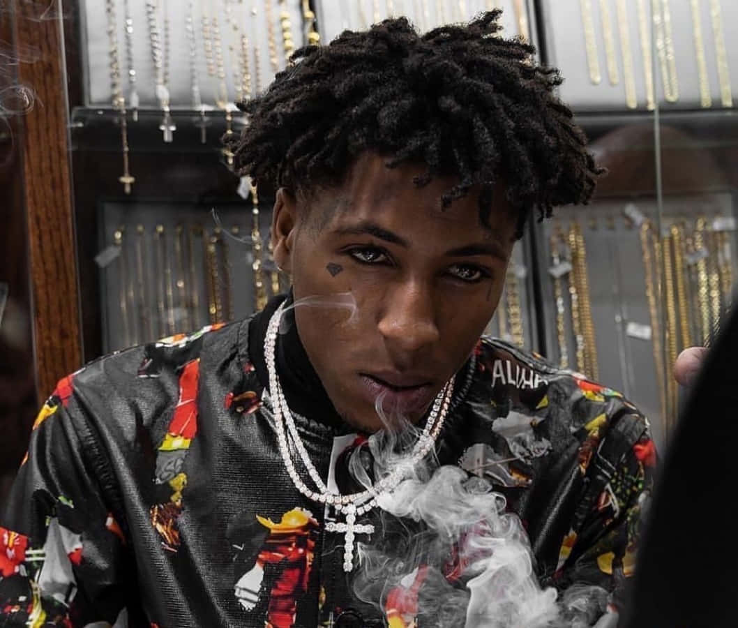 NBA YoungBoy 桌布 壁紙 — 一名年輕男子留著辮子，在珠寶店裡穿著色彩繽紛的夾克和鑽石鏈條，呼出煙霧。