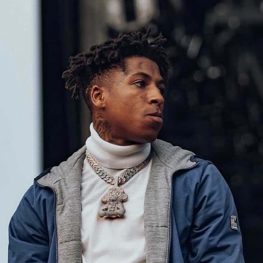 NBA YoungBoy 桌布 壁紙 — 一名年輕男子留著辮子，戴著金鏈吊墜，在模糊的城市背景下若有所思地凝視著。