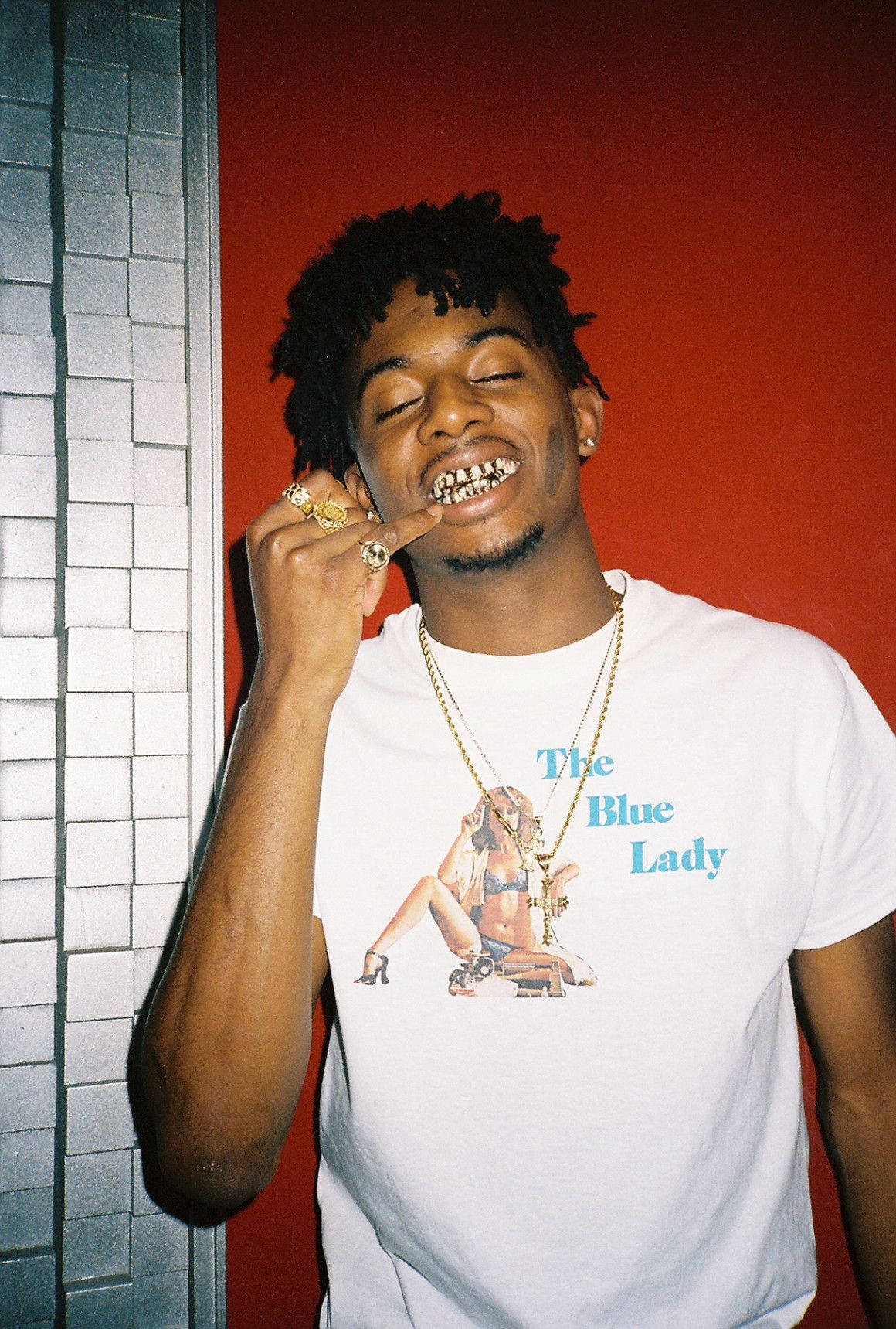Carti 桌布 壁紙 — 一名年輕男子留著辮子，露出金牙燦爛地微笑，同時將臉貼在紅牆和瓷磚柱上。