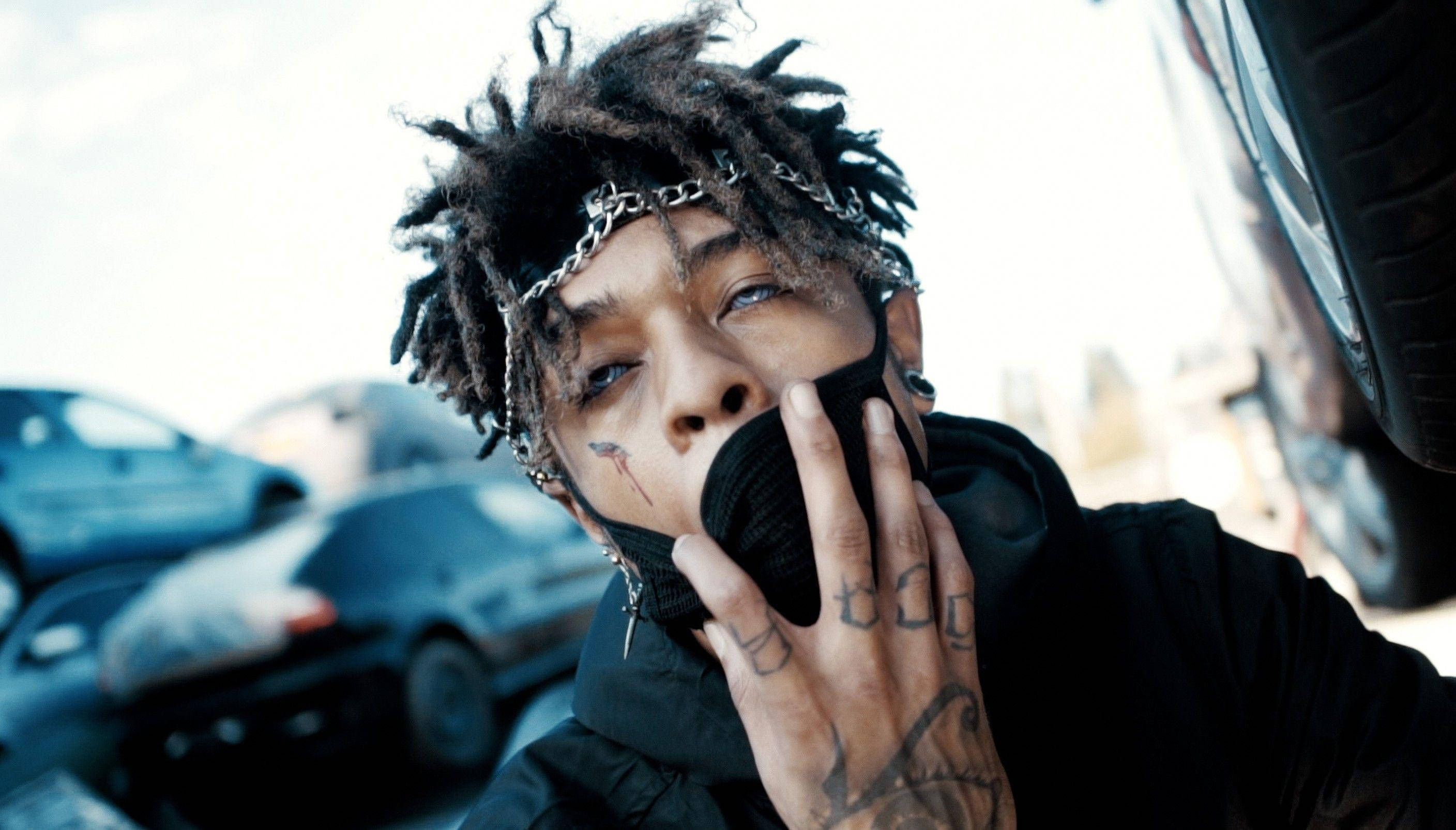 scarlxrd 桌布 壁紙 — 一名年輕男子留著辮子頭和刺青，在粗糙的城市環境中站在汽車附近，用黑色手套捂住嘴巴。